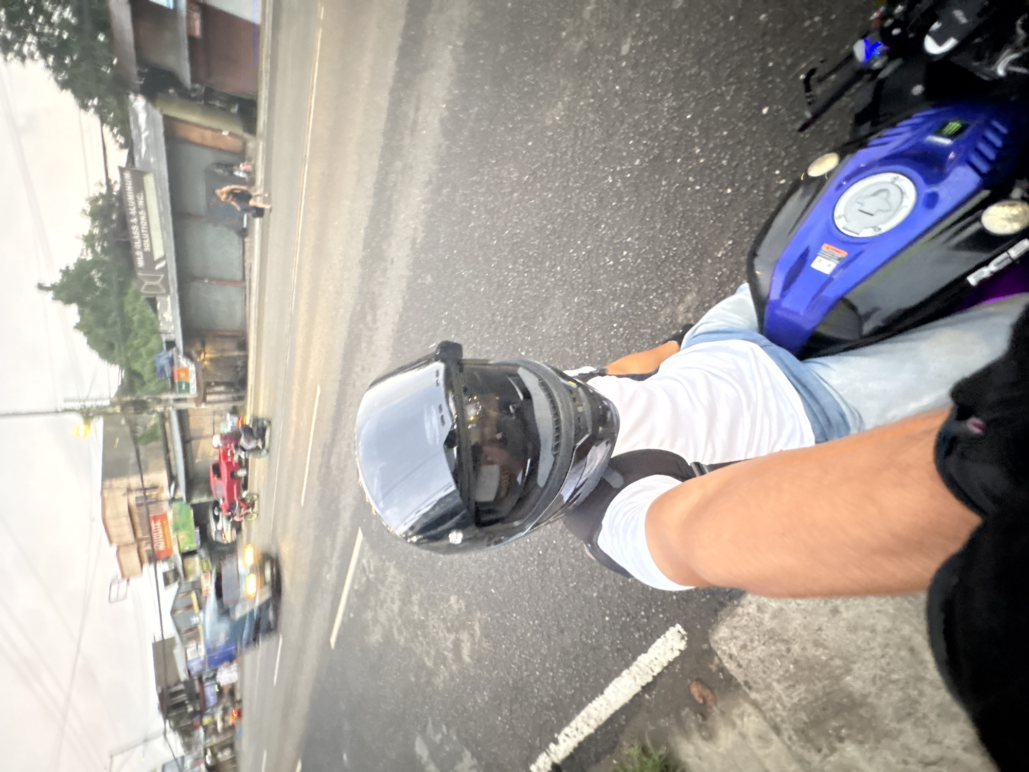 This is a first-person perspective photograph of a person riding a motorcycle on a street in Davao City, Philippines. The rider is wearing a black helmet with a tinted visor and a white t-shirt. The motorcycle is blue and black. The street is paved with asphalt and has white lane markings. In the background, there are buildings, a red pickup truck, a blue van, and other motorcycles. The sky is overcast, suggesting it is either early morning or late afternoon. The overall impression is that of a busy street scene in a Philippine city.