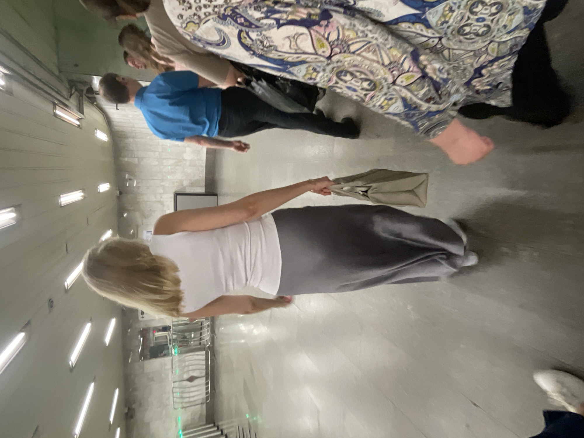 The image depicts an indoor environment, likely a metro station in Moscow, Russia, given the distinctive architectural style featuring illuminated ceiling panels and tiled walls. In the foreground, a woman with blonde hair, wearing a white sleeveless top and a long grey skirt, is walking away from the camera, carrying a beige tote bag in her right hand. Behind her, a man in a blue shirt and black pants is walking, and another individual in a patterned top is also visible. The ground appears to be a polished concrete or stone surface. Several silver turnstiles are visible in the background, suggesting a paid entry area. The lighting is bright and even, consistent with artificial illumination found in public transit stations.