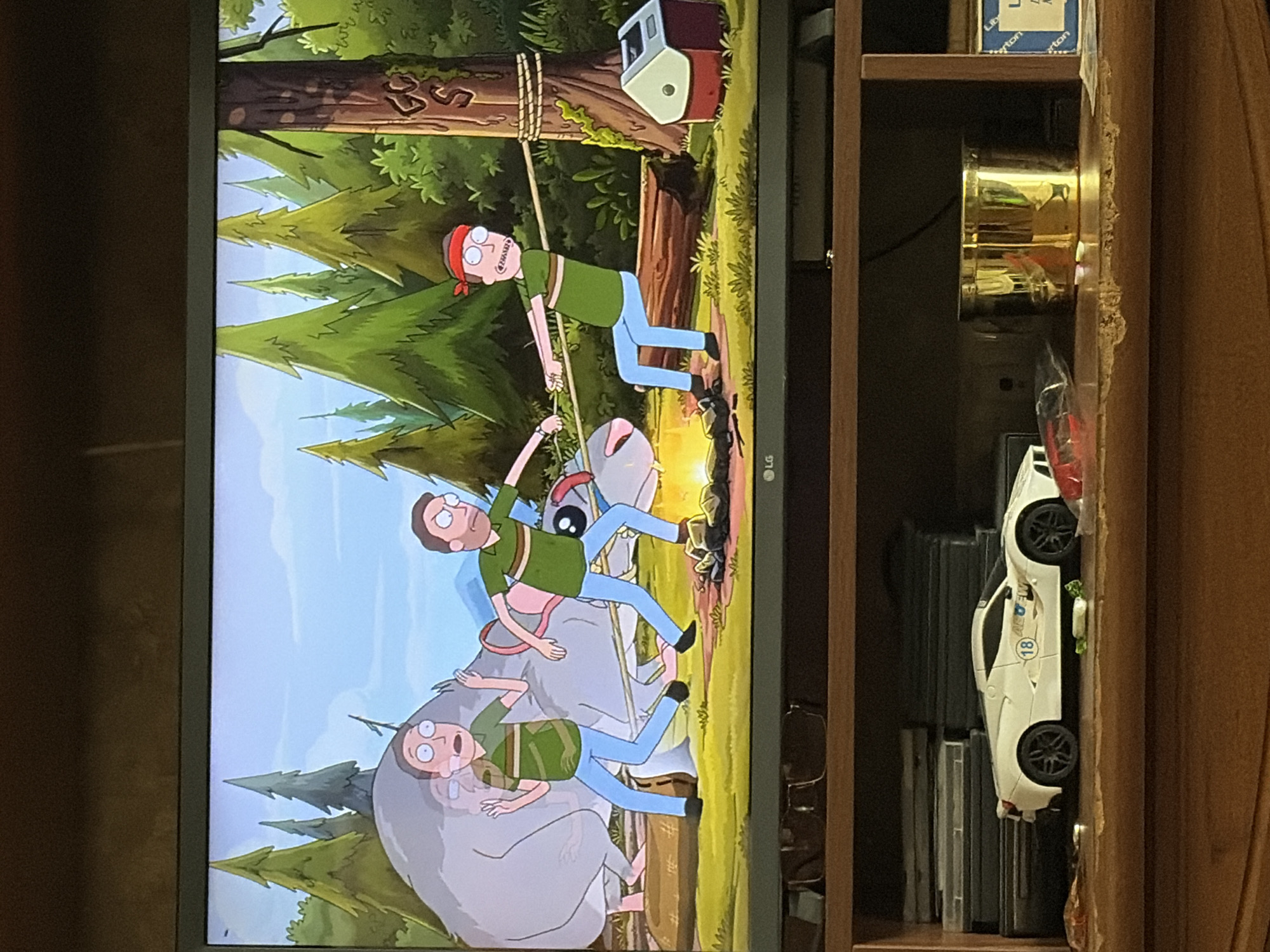 The image shows a television screen displaying a scene from the animated show Rick and Morty. The scene depicts two characters who appear to be Rick and Morty in a forest setting. One character, presumably Rick, is tied to a tree with a rope, while the other character, Morty, is pulling on the rope. A large, gray, rodent-like creature is also present in the scene, appearing to be a victim of some sort of predicament. In the foreground of the image, below the television, a white toy sports car is visible on a wooden shelf, along with a gold cylindrical container and stacks of what appear to be DVDs or game cases. The overall environment depicted on the television screen is a dense forest with tall pine trees and a grassy clearing.