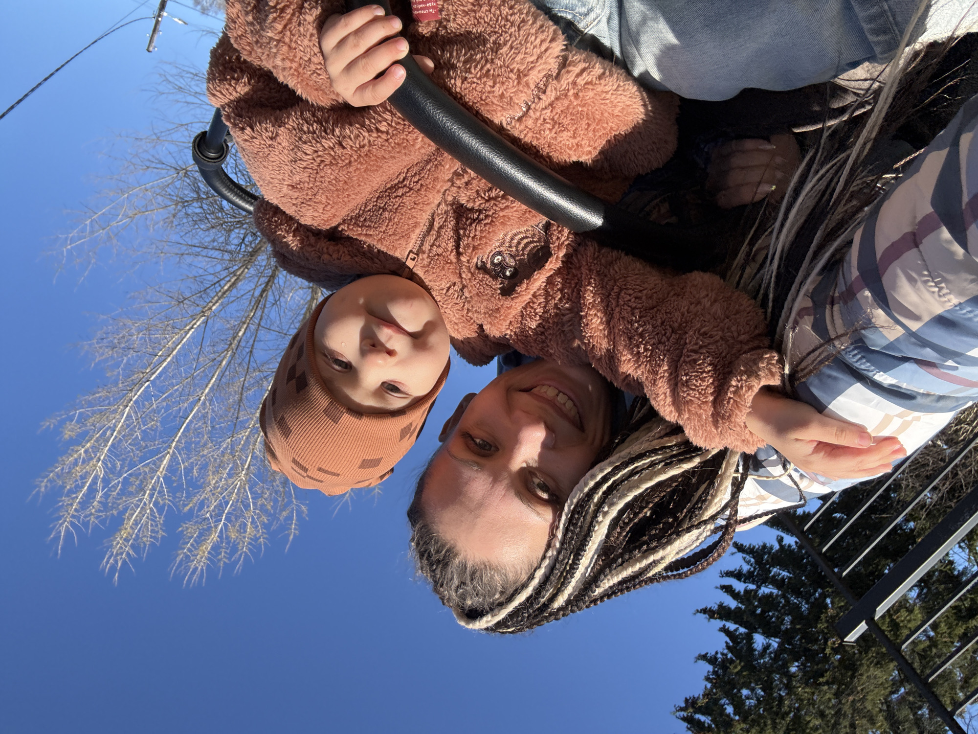 An overhead shot captures an adult and a young child looking down towards the camera from a low angle, set against a bright, clear blue sky. The adult, positioned on the right, has dark and light braided hair (dreadlocks style) and is smiling broadly with a cheerful expression. The child, on the left, wears a brown/orange ribbed knitted hat with a subtle checkered pattern and a fuzzy, light brown/tan jacket. Their face is partially visible, with eyes looking towards the camera.
A black, padded bar or handle is prominent in the foreground, held by the child's small hand, suggesting they might be in a stroller, swing, or carrier. The adult appears to be wearing a blue and white striped garment.
The background reveals bare tree branches against the vivid blue sky, indicating either late winter or early spring. In the distance on the far right, darker evergreen trees are visible, along with a black metal railing. A utility pole and power lines are discernible in the upper left portion of the image. The bright sunlight creates clear highlights on the subjects' clothing and faces. The overall mood is joyful and relaxed, likely an outdoor moment shared between the two individuals in Rusanovo, Russia. No discernible text is visible on any objects or clothing.