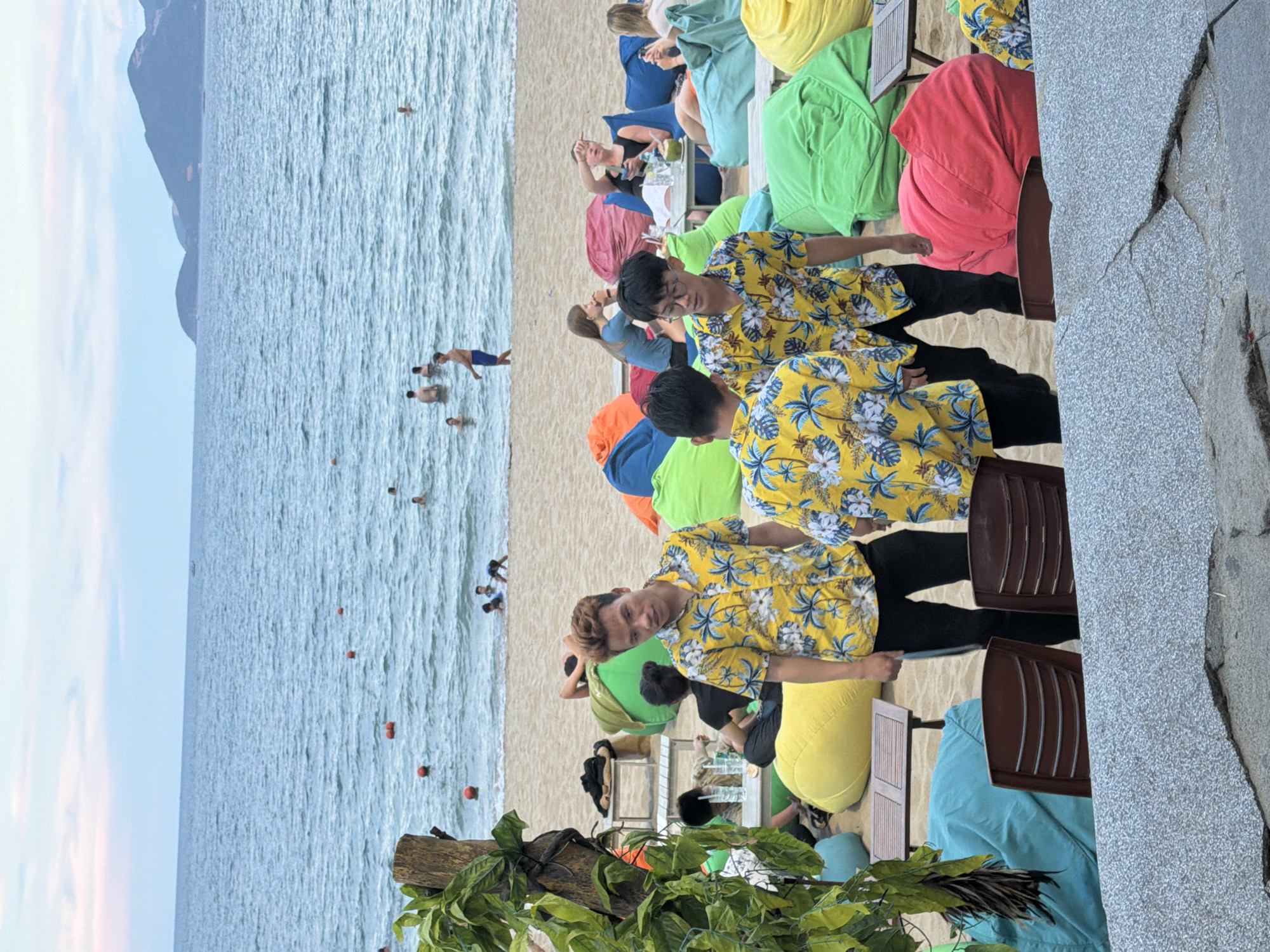 The image depicts a beach scene in Nha Trang, Vietnam, possibly in a beachside resort or restaurant area. In the foreground, three young men are standing, all wearing matching yellow floral print short-sleeved shirts and black pants. The man on the left, with reddish-brown hair, is looking directly at the camera with a neutral expression. The man in the middle, wearing glasses, is also looking towards the camera. The third man, partially visible on the right, is also wearing the same shirt.
Behind the men, the beach is furnished with numerous large, colorful beanbag chairs in red, yellow, green, and blue, arranged on the sand. Several small tables are interspersed among the beanbags. Further back, people are seen lounging on the beanbags and a few individuals are in the water. The ocean stretches to the horizon, with gentle waves and some buoys visible. In the distance, hilly islands are silhouetted against a pale sky. The overall atmosphere suggests a relaxed and casual beach environment.