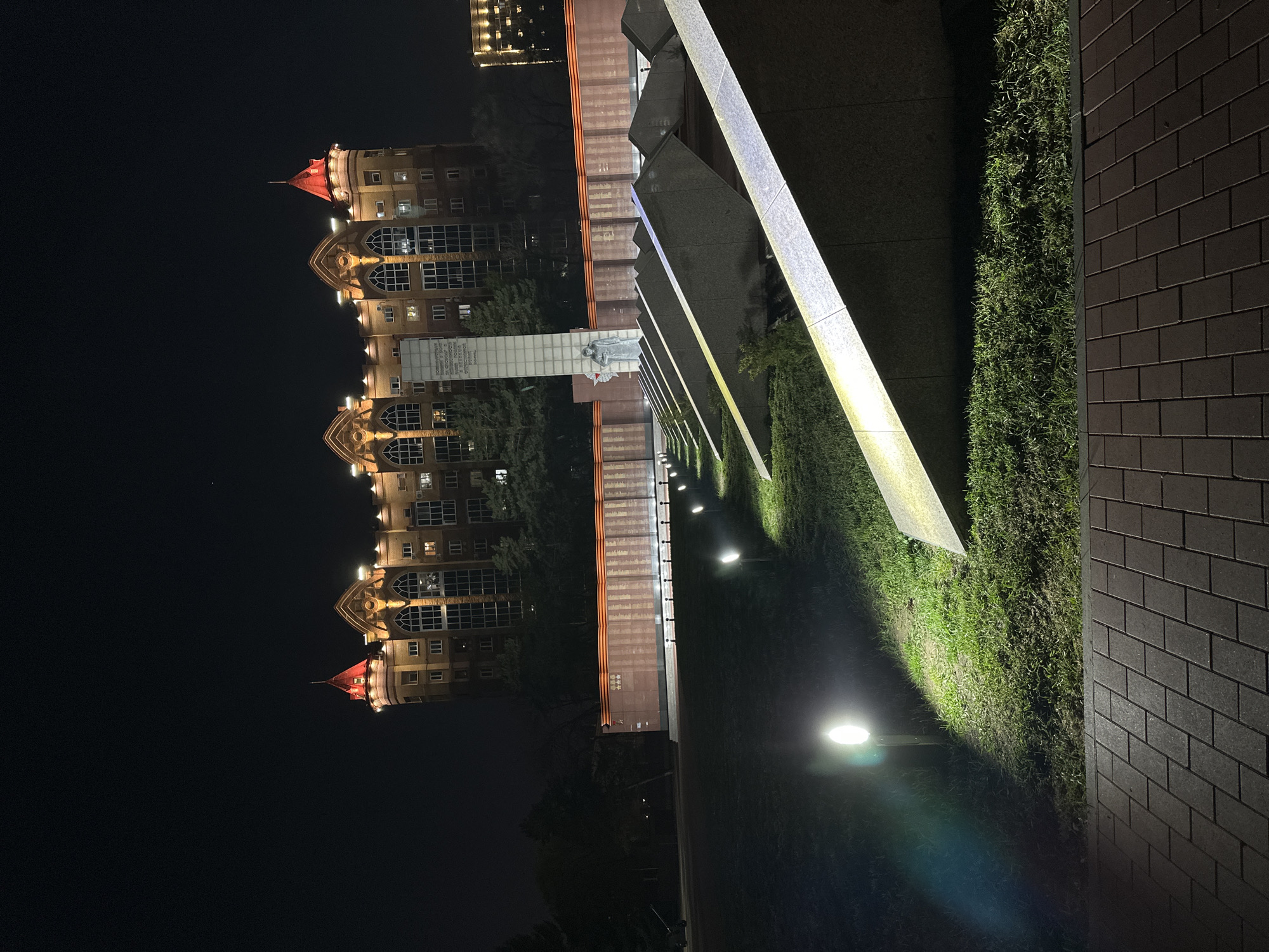The image shows a nighttime scene in Blagoveshchensk, Russia. A large, ornate building with red roofs is visible in the background. Closer to the viewer is a memorial complex, illuminated by spotlights. The complex consists of a series of gray structures and a central statue, which appears to be dedicated to war veterans. There's a neatly maintained lawn in front of the memorial, and a brick wall is visible on the right side of the frame. The scene is lit by streetlights and spotlights, creating a contrast between the illuminated memorial and the dark night sky.