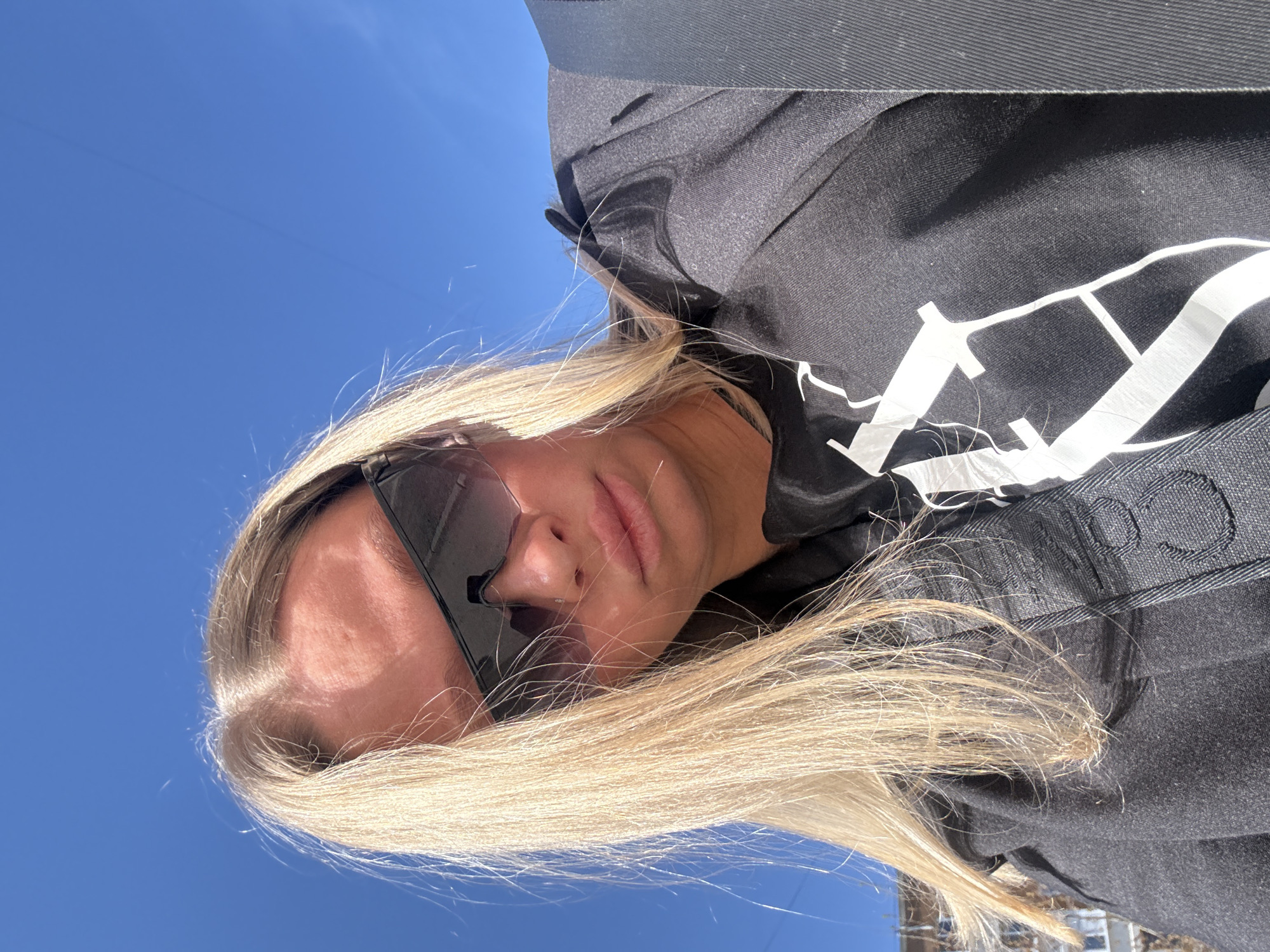 A woman with blonde hair is shown in a low-angle selfie, wearing sunglasses and a black jacket with a white design. The background is a clear blue sky. Part of a building is visible in the lower right corner. The location is possibly Perm, Russia.
