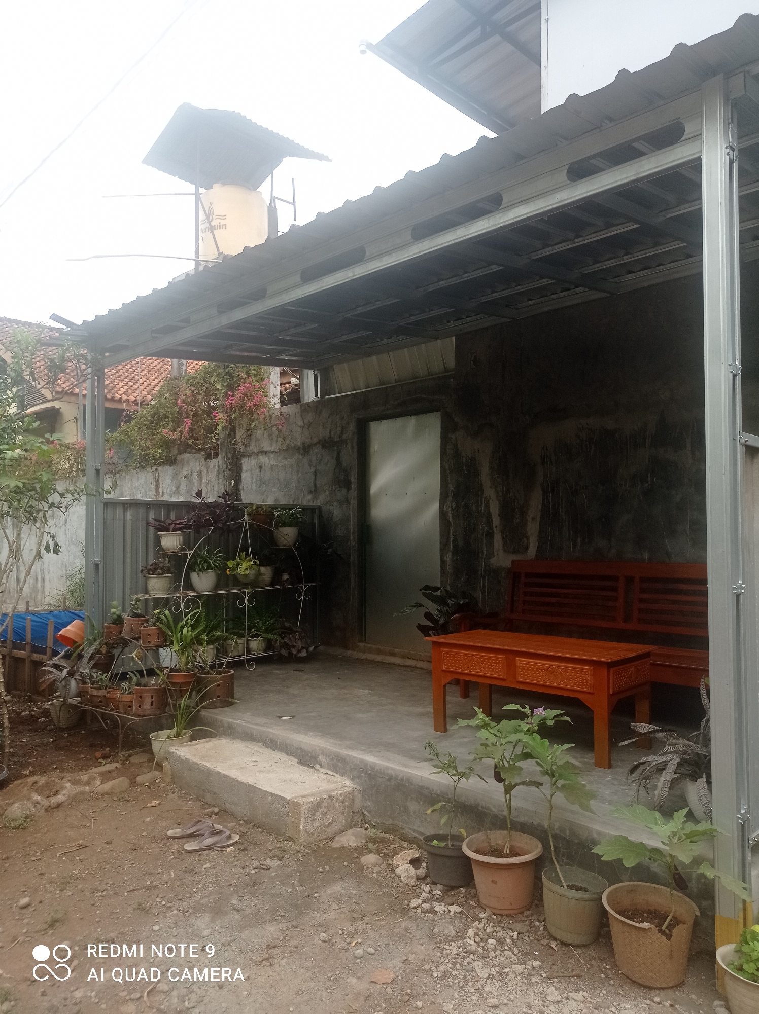 The image depicts an outdoor patio area, likely at a residence in Cipawon, Indonesia. The scene features a covered patio with a metal frame and corrugated roof. A wooden bench and a wooden coffee table are placed on the concrete floor of the patio. To the left, a multi-tiered metal plant stand holds various potted plants. Further to the left, a metal fence and a blue tarp are visible. In the background, a concrete wall with some greenery is present. Several potted plants are also situated on the ground in front of the patio. Overhead, a water tank on a metal structure is visible. The ground is a mix of dirt and gravel. No people are present in the image.