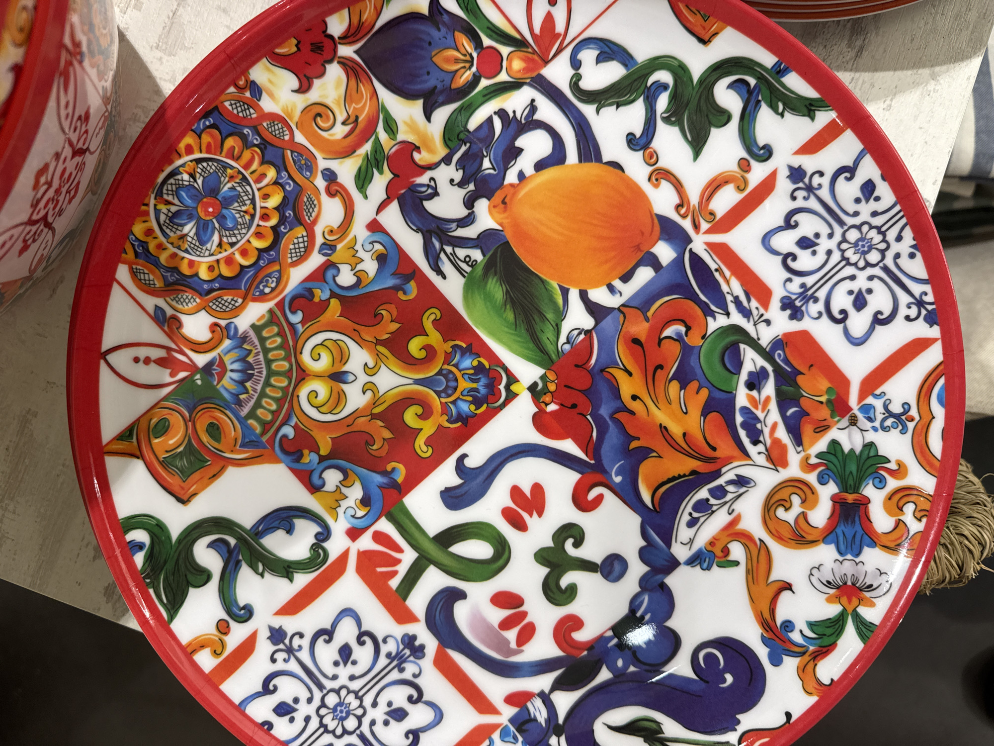 The image shows a brightly colored, round decorative plate with a red rim. The plate features a complex patchwork of designs, including floral patterns, geometric shapes, and a prominent image of an orange. The overall aesthetic is reminiscent of Mediterranean or Italian pottery. The plate is sitting on a surface, with a glimpse of a matching bowl visible in the upper left corner of the frame. The setting appears to be indoors. The image does not provide evidence to definitively place the location in Barcelona, Spain.