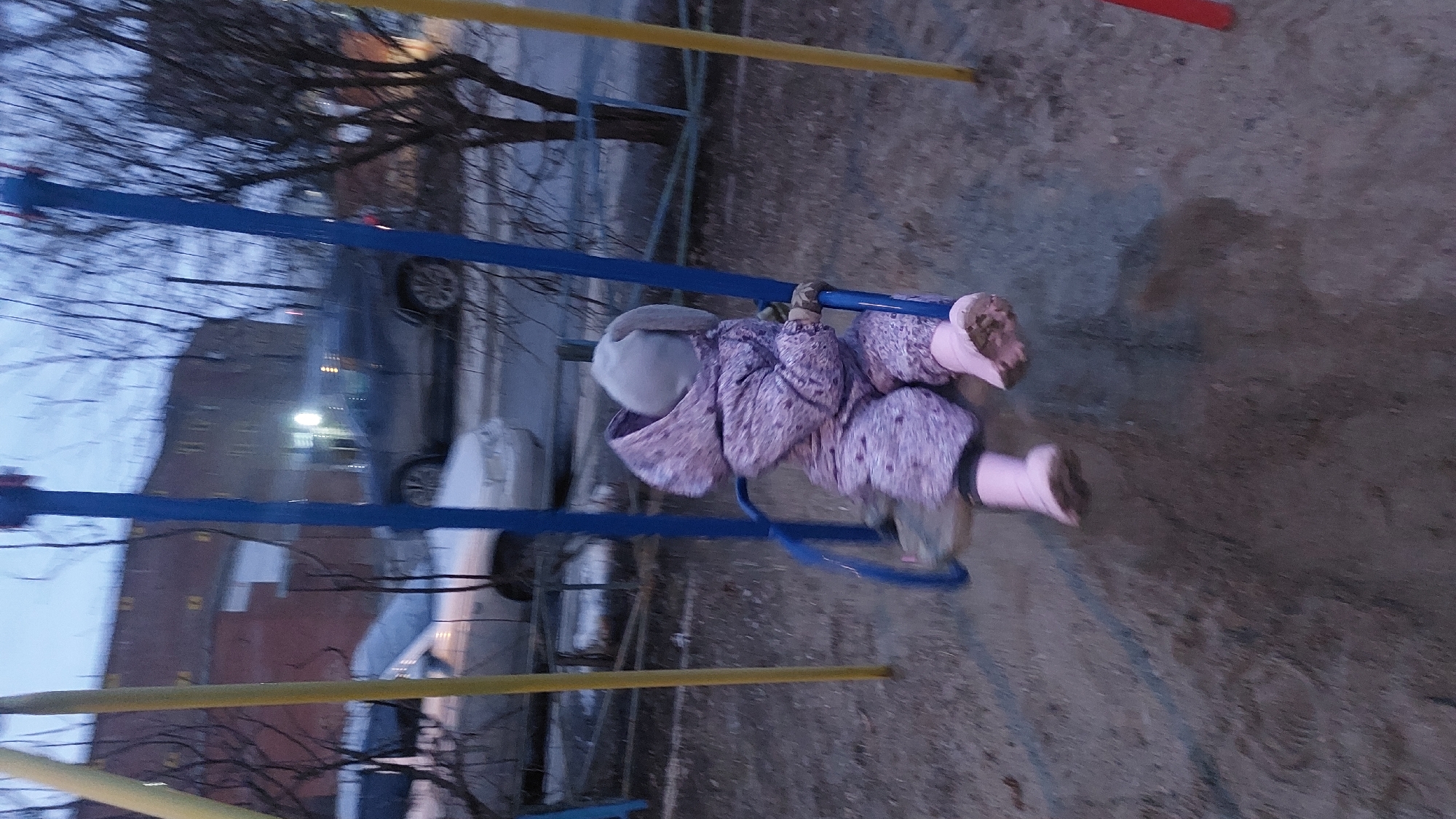 A child wearing a purple jacket, a hat, and pink boots is swinging on a blue swing set. The scene is outdoors, likely a playground, with bare trees in the background. There is a building and cars visible in the background, suggesting a city setting. It is dusk or early evening. Based on the location provided, the image may depict Novosibirsk, Russia.
