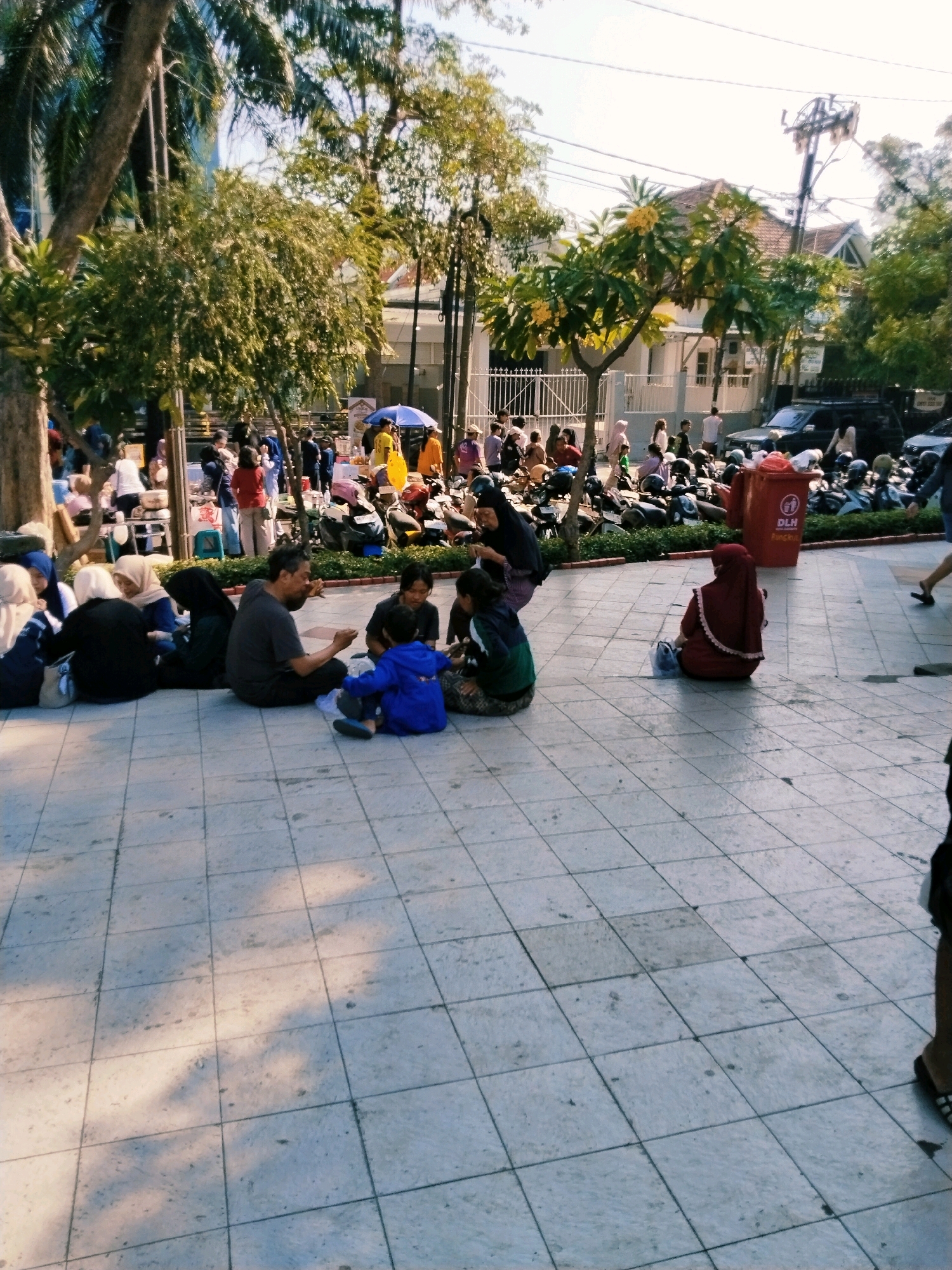 The image depicts an outdoor scene, likely in a public area of Surabaya, Indonesia, with a focus on a group of people sitting on a tiled ground. In the foreground, several individuals are seated in a semi-circle, some appearing to be engaged in conversation or eating. A man in a dark t-shirt is in the center, holding a piece of food or an object. To his left, a child in a blue jacket is also seated, and further left, other individuals are seated, some wearing hijabs. In the middle ground, there is a collection of motorcycles parked, and behind them, various stalls or vendors are set up. A blue umbrella is visible over one of the stalls. A red trash bin with a logo is also present. The background shows trees, buildings, and a utility pole with overhead wires, suggesting an urban environment. The lighting suggests it is daytime, possibly late afternoon, with shadows cast on the tiled surface.