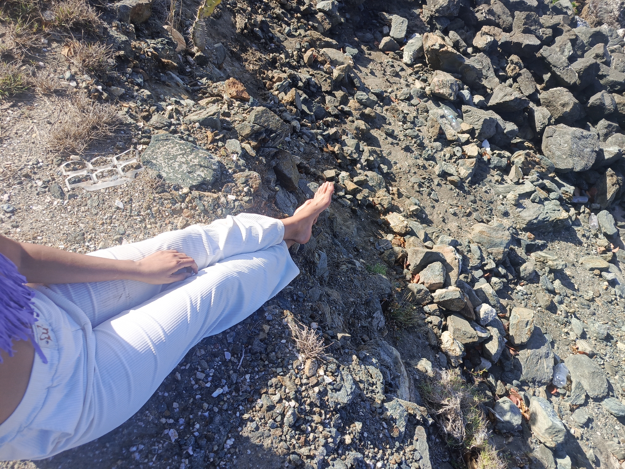The image shows a person's lower body and feet resting on a rocky, sun-drenched terrain. The person is wearing white, ribbed pants and a purple, fringed top, partially visible on the left side of the frame. Their right foot is bare and positioned on a flat patch of ground. The environment is characterized by an abundance of various-sized rocks and pebbles, with sparse dry vegetation scattered throughout. Some discarded plastic debris, including a six-pack ring and water bottles, are visible amongst the rocks, suggesting the location may be a natural area experiencing littering. The lighting suggests it is daytime with strong sunlight.