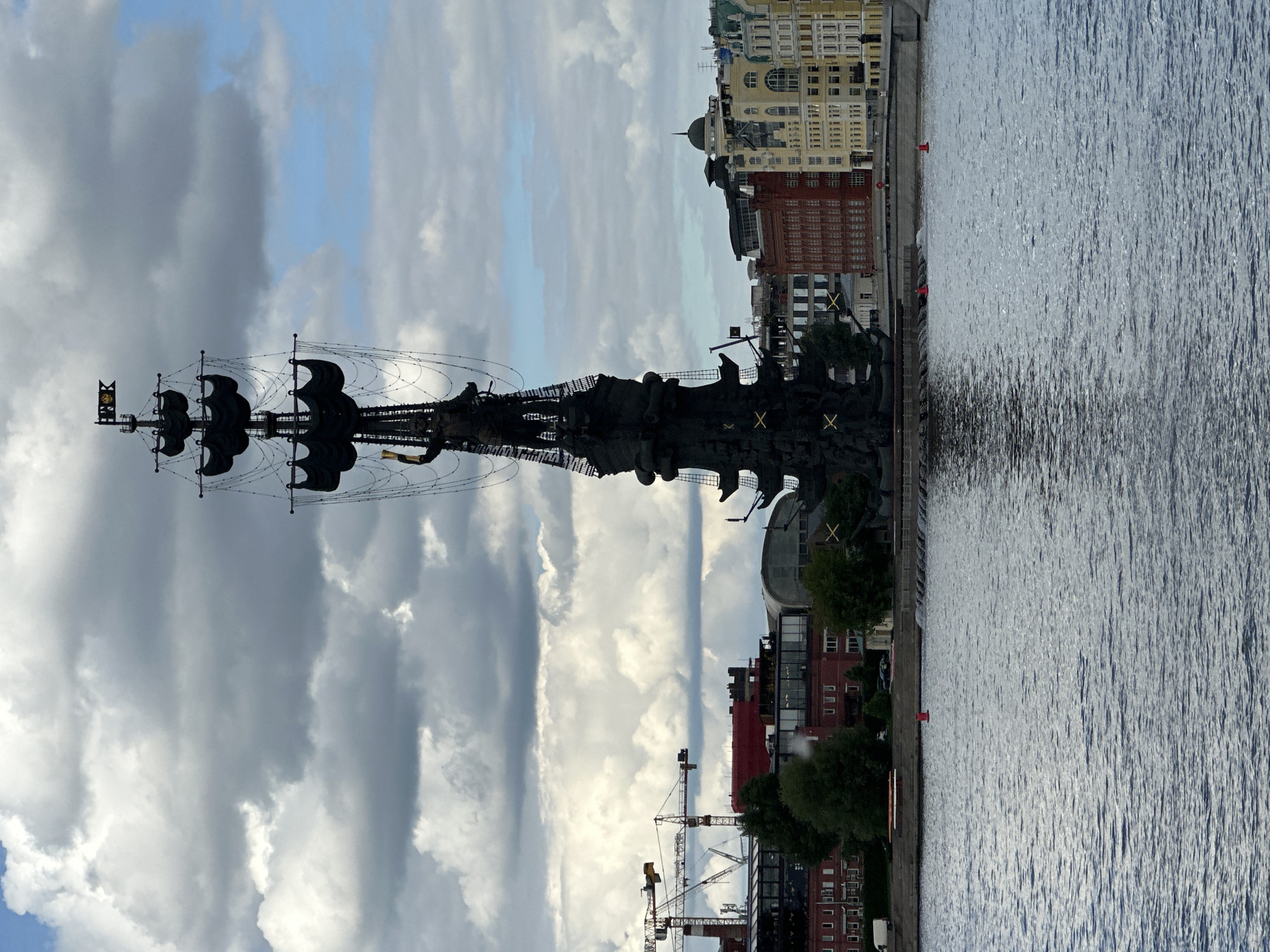 The image shows the Peter the Great Statue, a 98-meter monument to Peter I of Russia, located in Moscow, Russia, standing in the Moskva River. The statue depicts Peter the Great standing on a ship. The ship has masts and sails. There is a building line visible on the right side of the image. The water of the Moskva River is visible on the right and the sky with some clouds on the left. There are cranes visible on the background. The photo has been taken from a position that is in front of the statue.