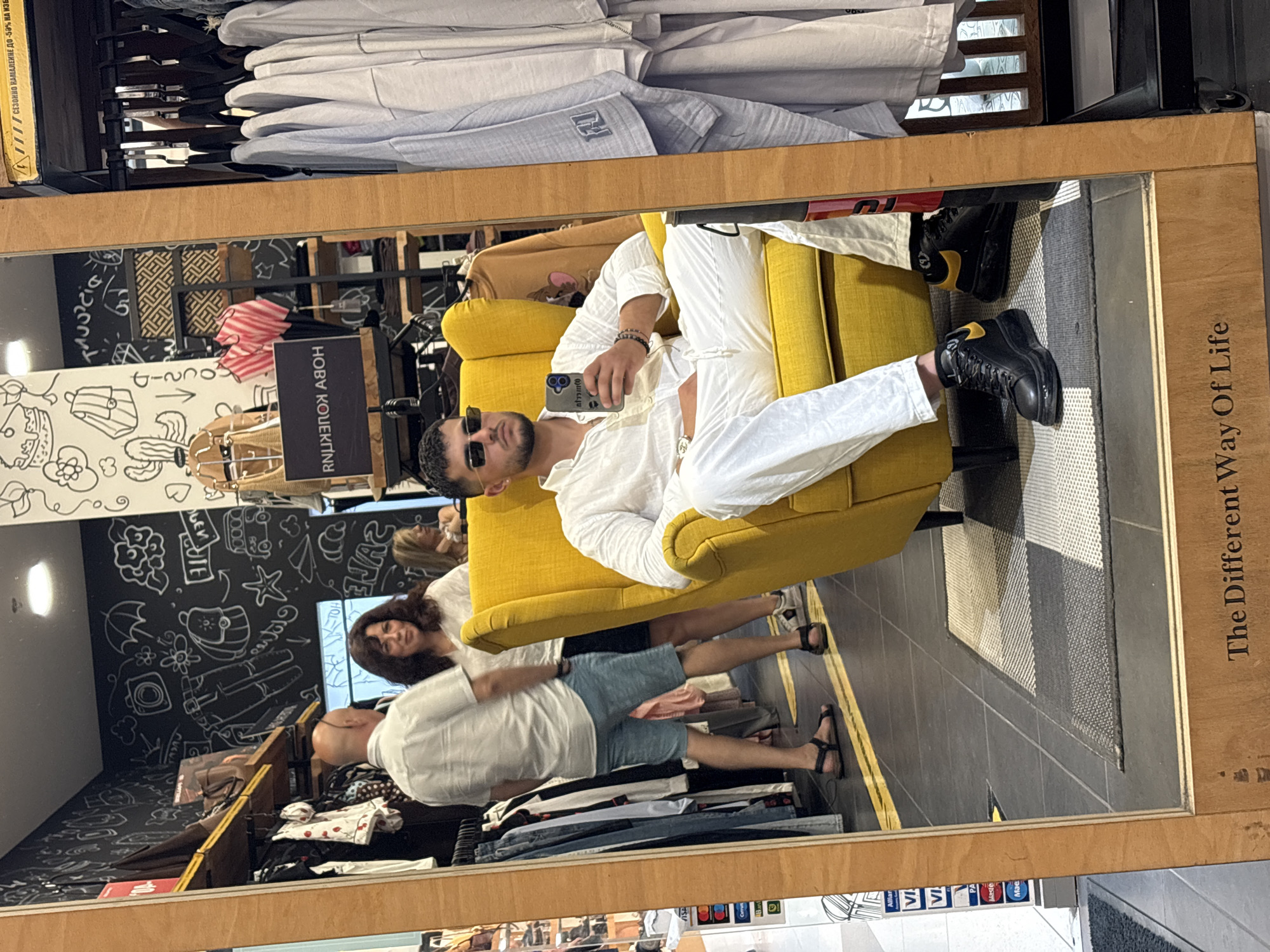 The image shows a reflection in a mirror of a man sitting in a yellow armchair and taking a selfie. He is wearing sunglasses, a white linen shirt, white linen pants, and black chunky sneakers with yellow accents. In the background, there is a clothing store with racks of clothes and a black wall decorated with white drawings. The mirror is framed in wood, and text reading "The Different Way Of Life" is visible at the bottom. The location is specified as a clothing store in the city of Ruse, Bulgaria.