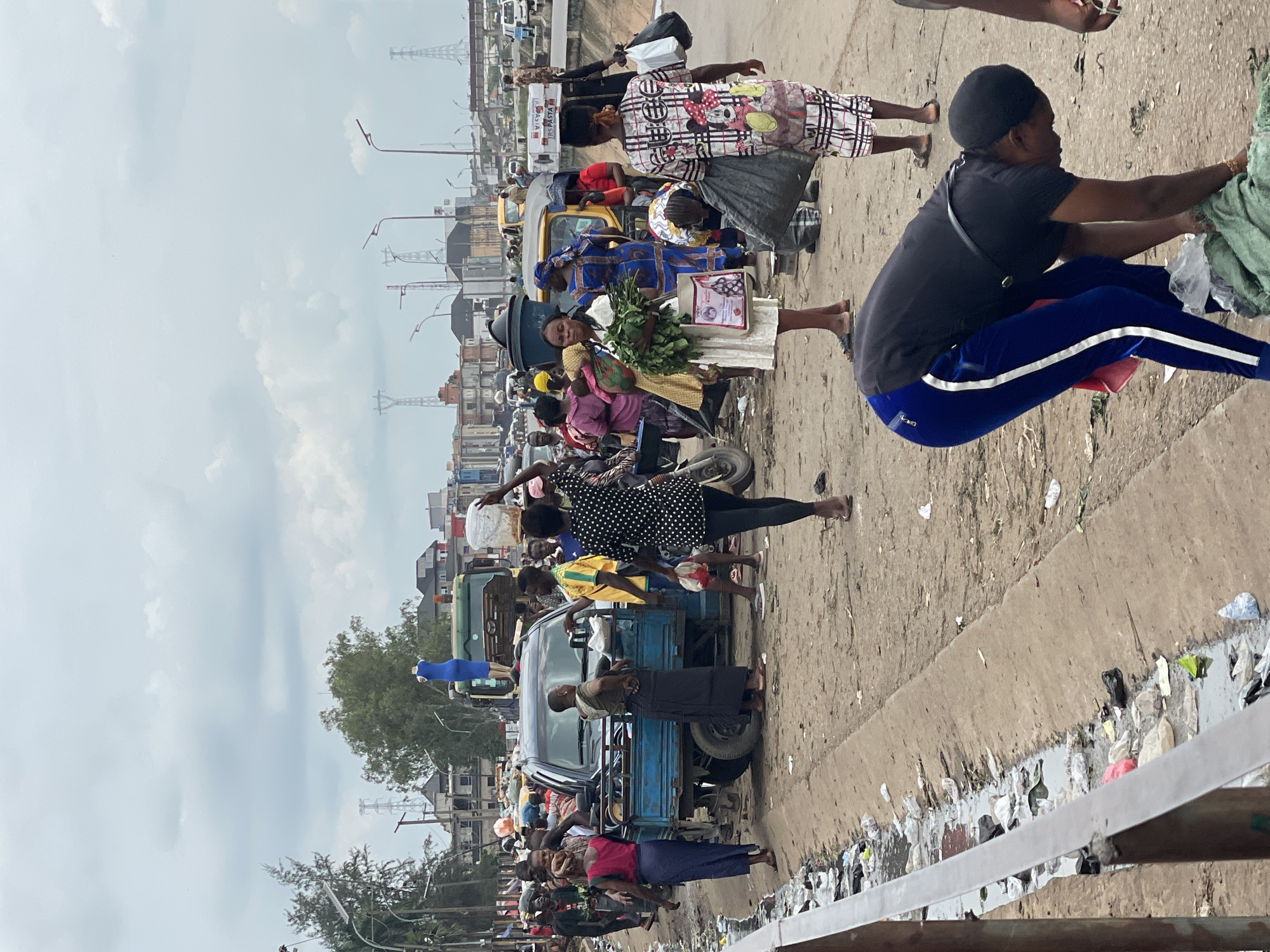 A street scene in Unknown, Unknown, shows a busy marketplace with many people and vehicles. The ground is littered with trash, and a narrow ditch filled with water and debris runs along the side of the road. In the foreground, a woman in a black shirt and blue pants bends over to pick up something from the ground. Behind her, a woman in a black and white polka dot dress walks away from the camera. A blue pickup truck is parked in the middle of the scene, with several people standing around it. A mannequin dressed in a blue gown stands on top of a green truck in the background. The sky is overcast with clouds.