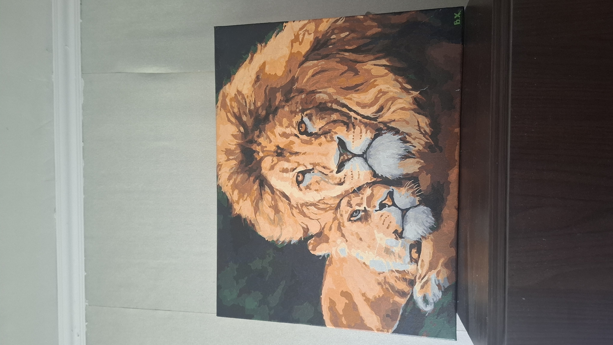 The image displays an acrylic painting of two lions resting on a dark brown wooden surface, positioned against a light-colored wall with white crown molding. The painting features a large male lion with a prominent orange and brown mane in the foreground, looking directly forward with orange eyes. A female lioness is depicted to the left, her head resting near the male lion's shoulder, with her gaze directed downwards. Both lions are rendered in various shades of orange, brown, grey, and white, against a dark green and black background. The signature "B.X." is visible in green in the bottom right corner of the painting. The dark brown wooden surface supporting the painting exhibits a horizontal raised trim element. The wall behind the painting is light grey or beige with subtle vertical textures, beneath a white crown molding that separates it from a white ceiling. The scene is illuminated by ambient interior light.