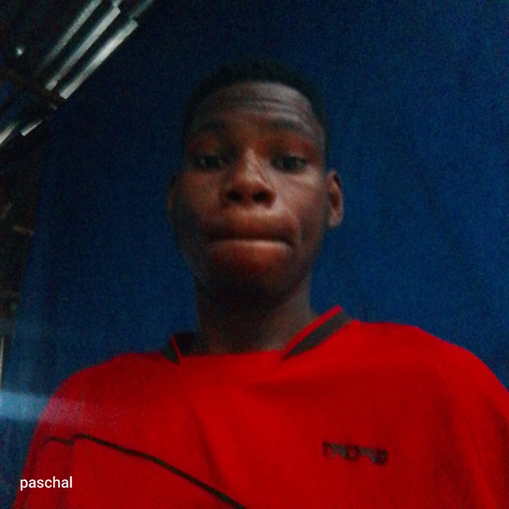 The image is a head-and-shoulders selfie of a young individual, appearing male, with dark skin and short dark hair. The person is centered in the frame, looking directly at the camera with a neutral expression and slightly pursed lips.
They are wearing a red short-sleeved shirt with black trim on the collar and shoulders, and a black diagonal line across the chest. A small, dark, and blurry logo or text is visible on the right side of the shirt's chest area, but it is illegible due to blurriness.
The background is a plain, dark blue surface, likely a wall. In the upper left corner, several light-colored, metallic, and somewhat blurry parallel bars or slats are visible, possibly part of a ceiling structure or fixture. The overall lighting is dim, suggesting an indoor setting with artificial light.
In the bottom left corner of the main image, the word "paschal" is visible in white text. The image quality is somewhat grainy due to the low light.