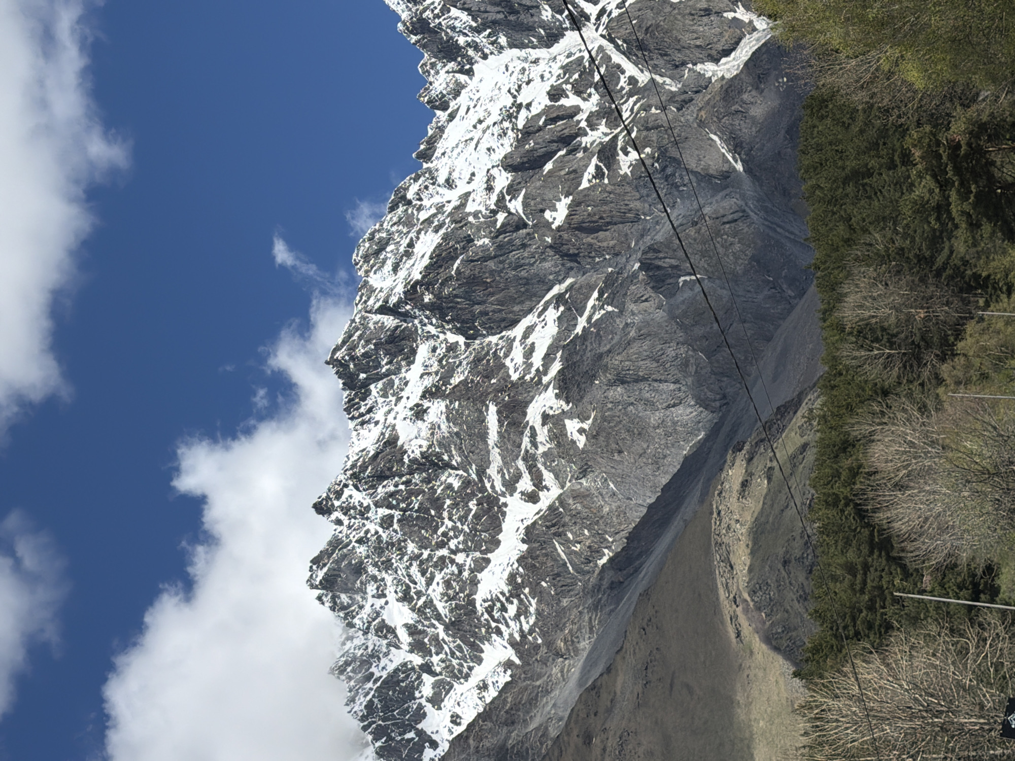 The image depicts a steep, snow-capped mountain located near Stepantsminda, Georgia. A dark, rocky face makes up a large portion of the mountain's structure, with patches of snow clinging to its rugged surface. Overhead power lines diagonally cross the mountainside. To the right of the mountain, a forested area merges with drier, less vegetated slopes. In the background, a bright blue sky is partially obscured by fluffy white clouds.