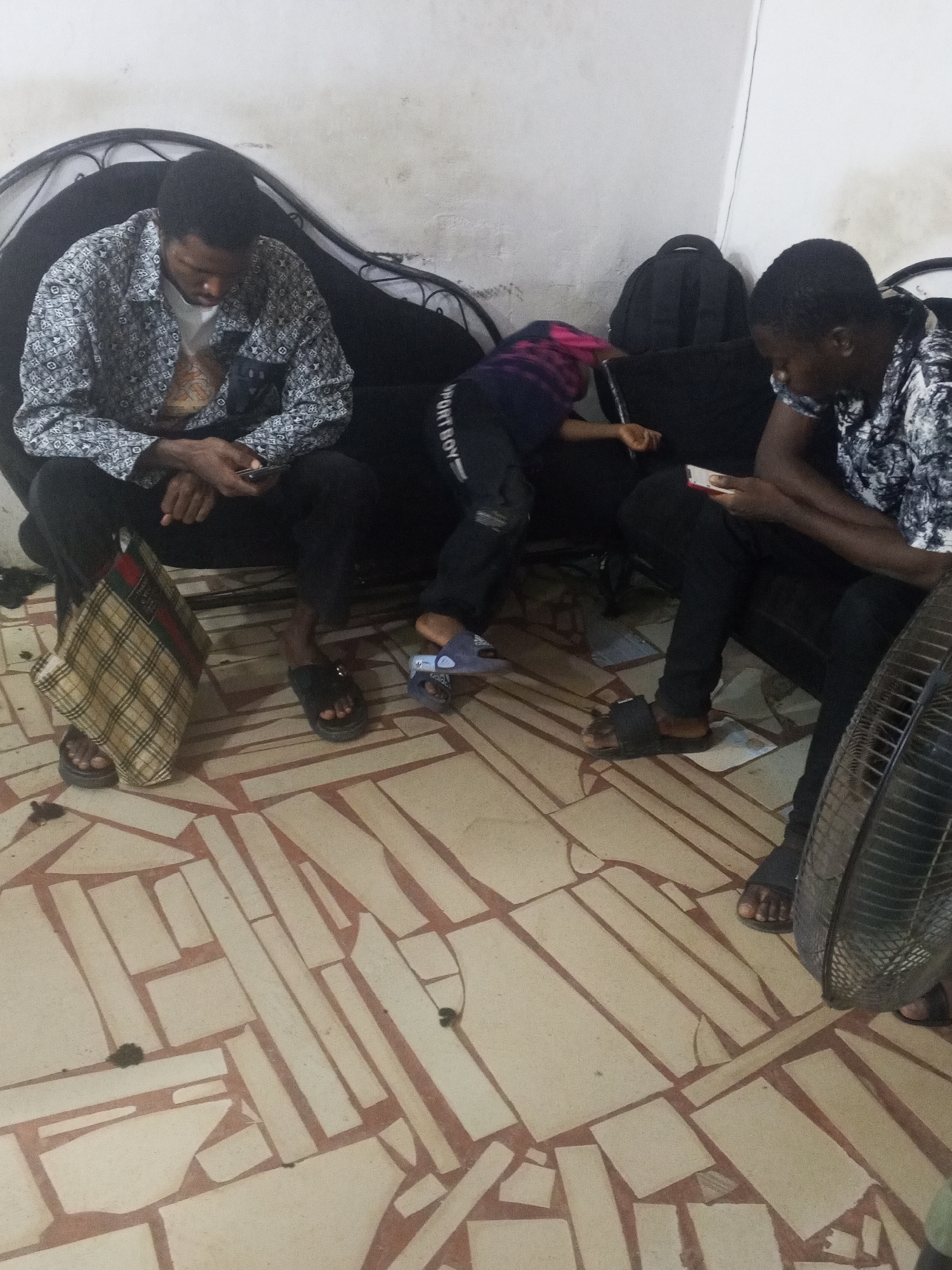 Three people are on a black couch in an indoor location with a patterned tile floor. The person on the left wears a patterned shirt, black pants, and sandals and is looking at a phone. The middle person wears dark clothing with "SPORT BOY" visible on the pant leg and a pink shirt and is lying horizontally. The person on the right wears a patterned shirt, black pants, and sandals and is looking at a phone. A checkered bag sits on the floor next to the first person. A black backpack is behind the middle person. A fan is visible on the right. Debris is scattered on the floor. The location is Gwong, Nigeria.