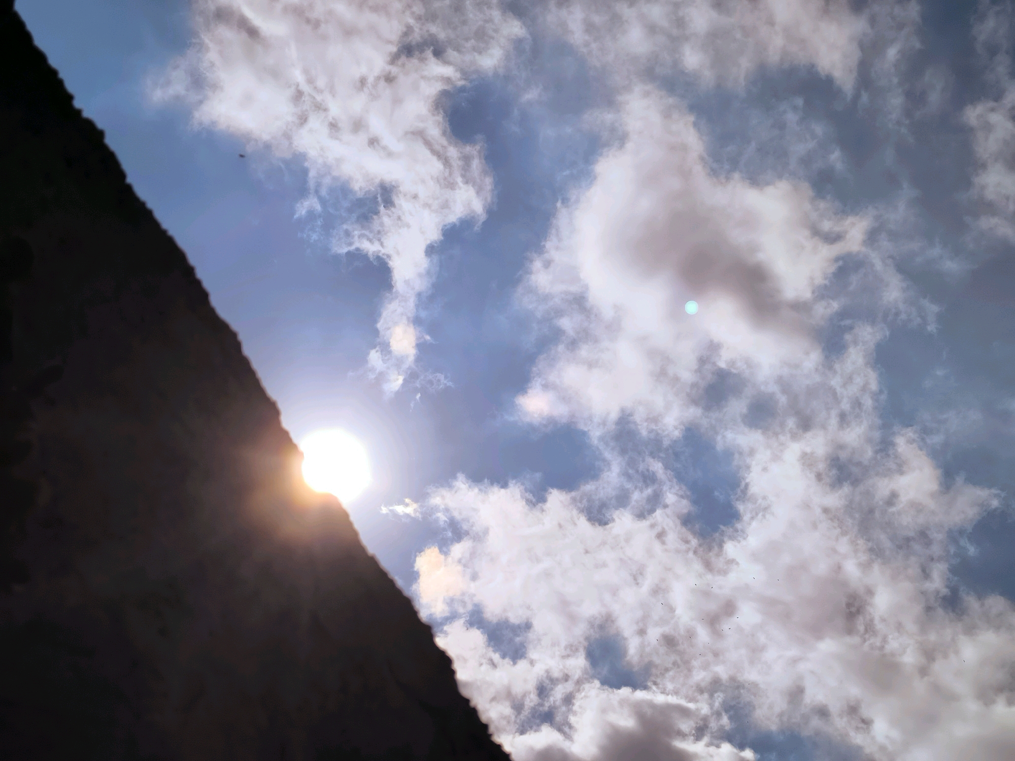 The image shows a low-angle shot of a cloudy sky with the sun peeking out from behind a dark, jagged edge on the left side of the frame. The clouds are a mix of white and gray, with some appearing wispy and others more dense. The sun is bright and casts a warm glow, creating lens flare. There are several small, dark objects scattered across the sky, which appear to be birds. The overall scene is one of a dramatic sky with the sun and clouds in focus, framed by an undefined dark object. The location is not identifiable from the image itself.
