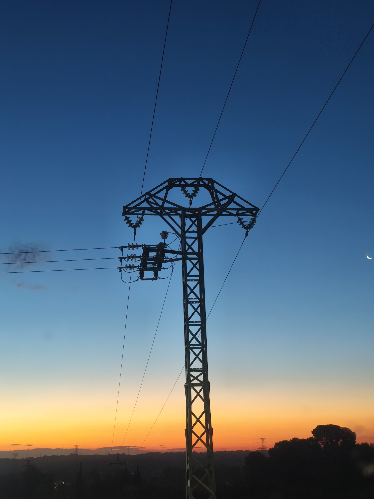 The image shows a tall, steel electrical transmission tower silhouetted against a gradient sky, transitioning from orange near the horizon to deep blue overhead. Power lines stretch from the tower towards the viewer and to the right. The tower is located in Riba-roja de Túria, Spain, as suggested by the landscape and architectural features visible in the background. In the lower portion of the frame, silhouetted trees and distant structures can be seen along the horizon. A crescent moon is visible in the upper right.