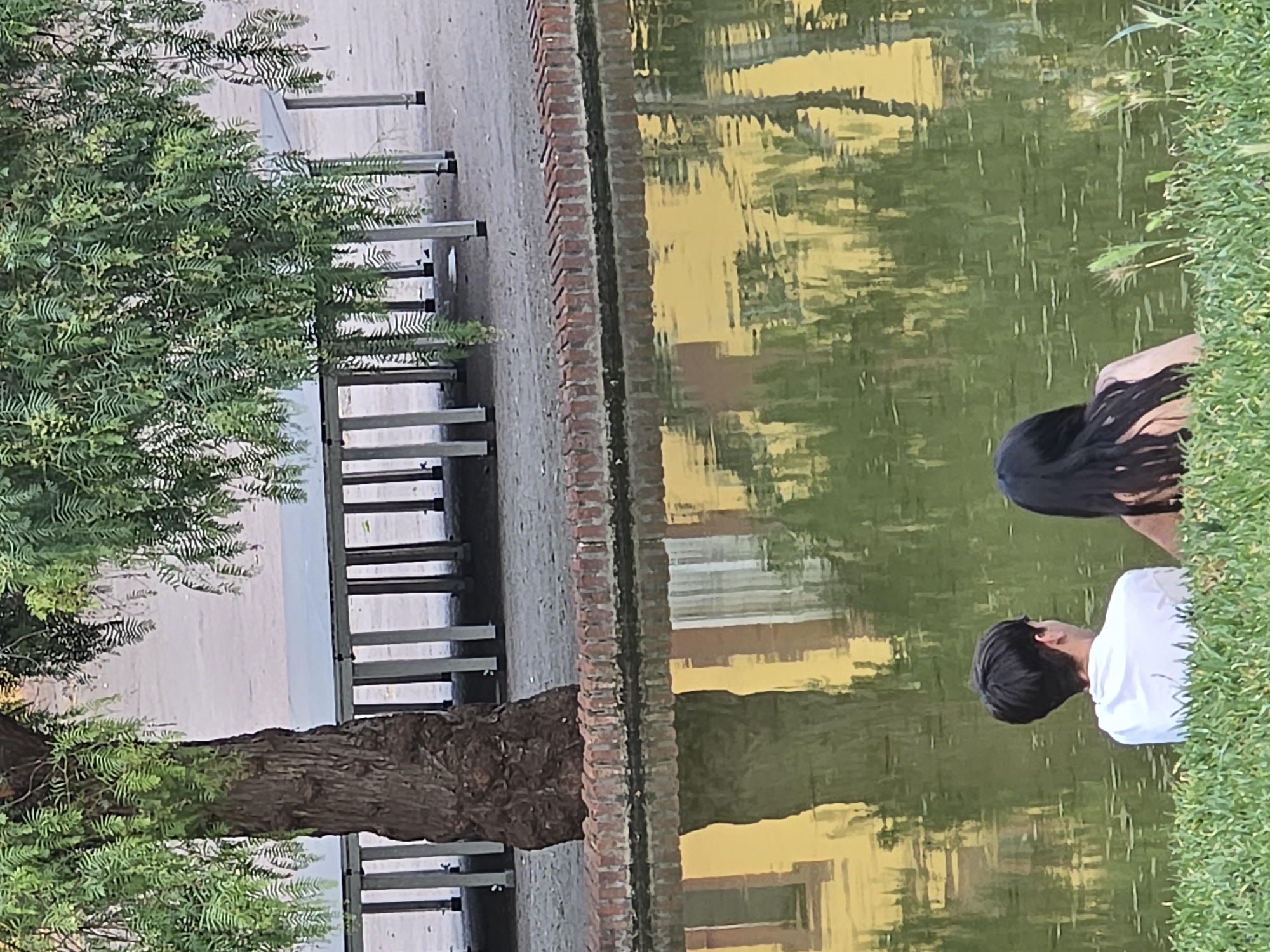 In Cornellà de Llobregat, Spain, two individuals, one with short dark hair wearing a white t-shirt and another with long dark hair, sit facing a pond's reflective surface. The pond water displays a distorted yellow building and tree reflection. The foreground features lush green grass, while the pond is bordered by a brick wall. In the background, a tree with dense green foliage partially obscures a row of white tables supported by metallic legs. The ground appears to be a sandy or gravelly surface.
