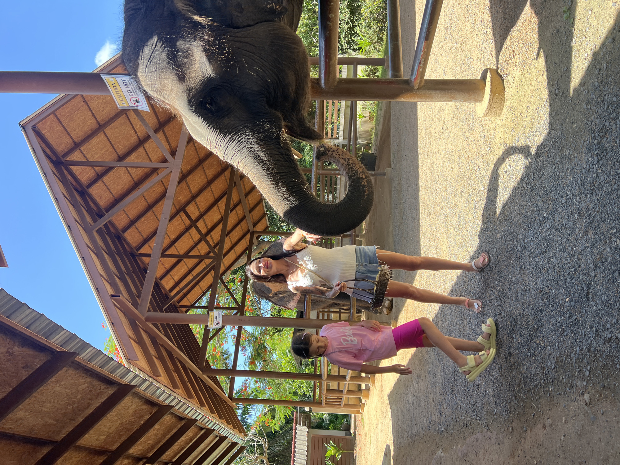 A photograph taken in Baan Thurian, Thailand, shows a partial view of an elephant with a woman and a young girl standing underneath it. The elephant is mostly out of view, with its head and trunk prominently visible. The woman is wearing a white top and denim shorts and is positioned behind the elephant. The girl is wearing a pink top and pink shorts and stands closer to the foreground. They appear to be interacting with the elephant, as the trunk extends down towards them. They are standing on a gravel surface under a wooden structure. In the background, lush tropical foliage is visible.