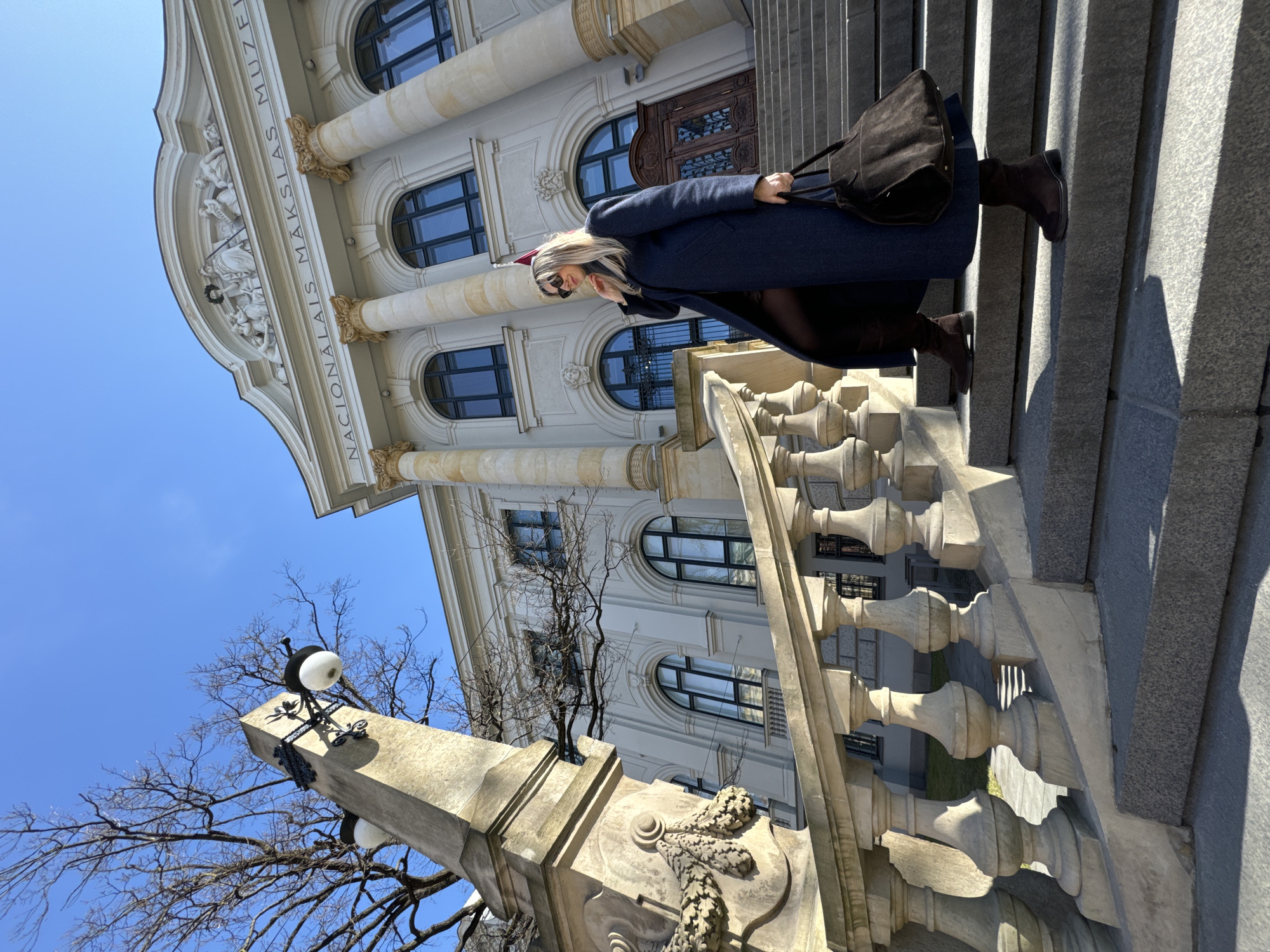 A woman wearing a navy blue coat, dark tights, and brown boots walks down the stairs of the Latvian National Museum of Art in Riga. She carries a large brown bag over her shoulder. The ornate facade of the museum is visible, with its columns, sculptures, and arched windows. A stone balustrade lines the stairs, and a lamp post stands in the foreground. The sky is blue and clear. The museum's name, "NACIONALAIS MAKSALAS MUZEJS," is inscribed on the building.