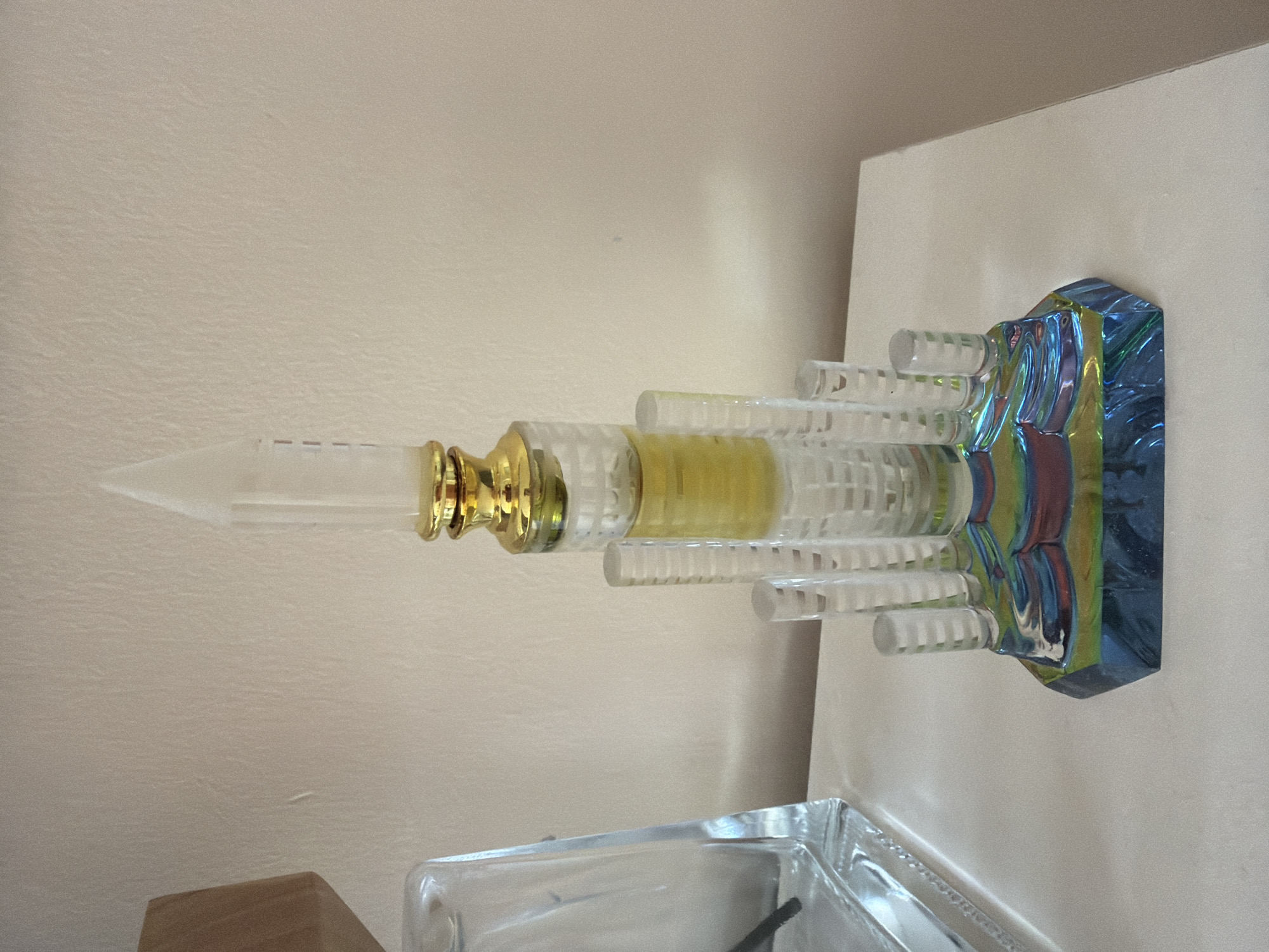 The image features a striking decorative object, resembling a modern skyscraper or monument, set against a plain, light-colored background. The object stands upright on a light-toned, flat surface, likely a table or shelf.
The main subject is a tall, slender structure primarily composed of clear, transparent material, possibly acrylic or glass. It tapers upwards to a pointed tip. A prominent golden-colored section encircles the middle portion of the object. Inside the transparent structure, a yellow substance, appearing as either a liquid or a solid element, is visible. Multiple clear, cylindrical rods or extensions protrude horizontally from the sides of the central column at varying levels, enhancing its architectural design.
The base of the object is distinctive, showcasing an iridescent or holographic finish that reflects a spectrum of colors, including vibrant blues, greens, purples, and hints of red, creating a dynamic visual effect. The ambient lighting suggests an indoor setting, likely during the daytime, casting soft shadows on the pale wall behind the object.
In the lower part of the frame, another clear glass or acrylic object is partially visible, appearing to be a container or a complementary decorative item. There are no people present, and no specific activity is taking place. The scene is a static still life. No text is discernible within the image. While the location context is Chervena voda, Bulgaria, no specific visual cues within the image itself indicate this location.