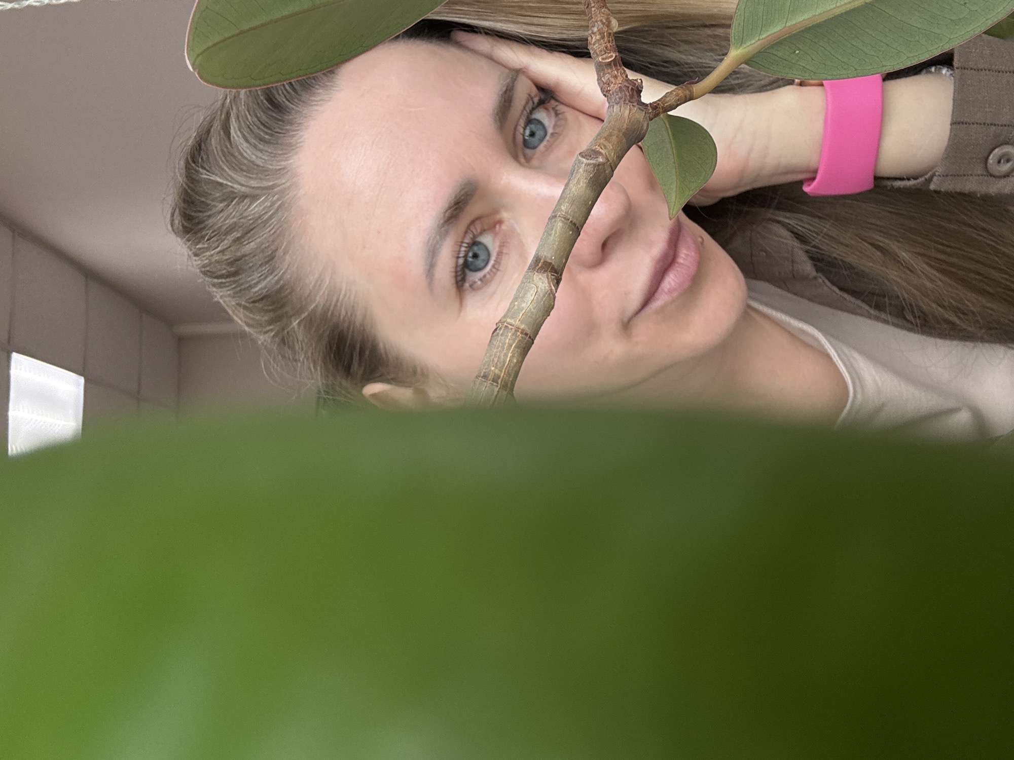 The image depicts a woman with fair skin and blue eyes gazing upwards, her gaze partially obscured by the stem and leaves of a green plant in the foreground. She rests her head on her hand, which is adorned with a pink fitness tracker on her wrist. She is wearing a light-colored top and a brown shirt or jacket. Her hair is light brown with visible graying strands. The background consists of a light-colored wall and a glimpse of a tiled surface. The location is not discernible from the visual information in the provided image, and the presence of the city of Chaikovsky, Russia cannot be determined without external information or explicit visual clues within the image itself.