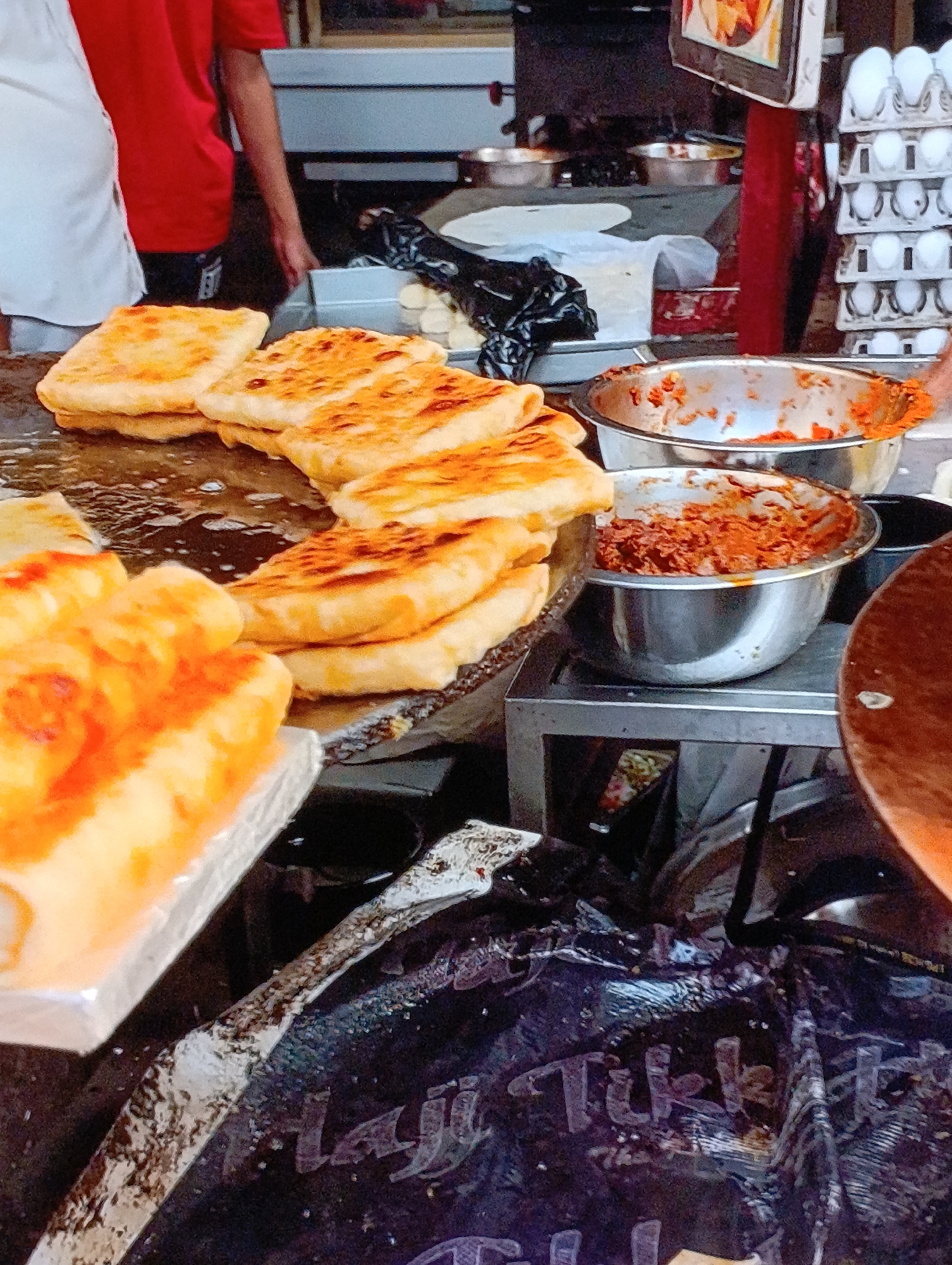 The image depicts a street food vendor preparing and selling food in Unknown, Unknown. There are two people visible, one wearing a red shirt and the other wearing a white shirt, implying they are likely vendors or assisting. The main subject of the image is food: several square, golden-brown, likely cooked, flatbreads are prominently displayed on a cooking surface, surrounded by a shimmering oily or buttery liquid. There are also two metal bowls containing filling, the contents of which appears to be orange or reddish in color, likely a spicy vegetable or meat mixture, or sauce. In the background, several uncooked flatbreads are visible, covered with plastic bags, and containers with eggs. The overall environment suggests a busy food stall, where food preparation is occurring, which is an activity that can be inferred from the subjects, food, and cooking surfaces.