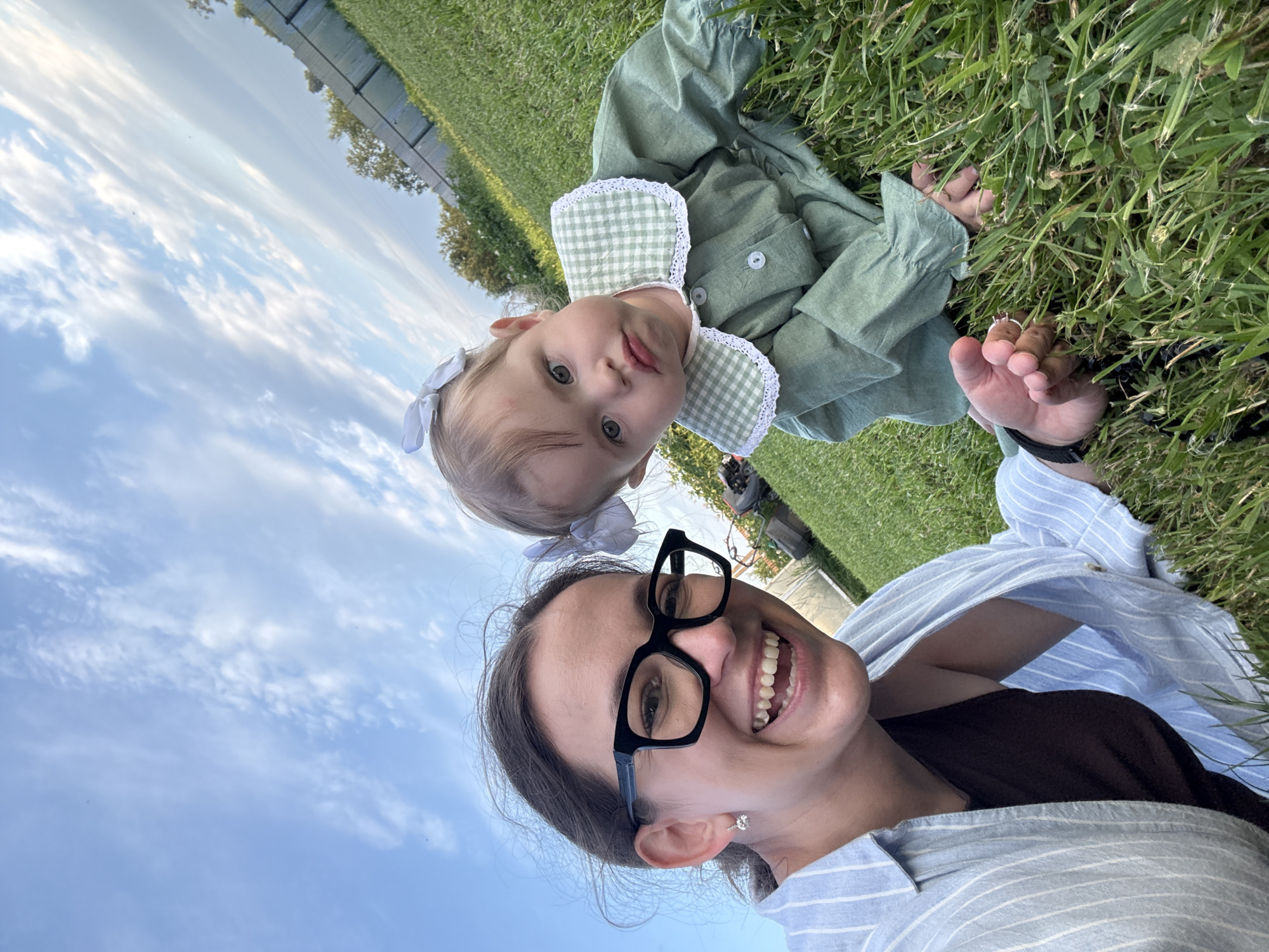 The image shows a woman with black-rimmed glasses smiling widely, and a young child with a white bow in her hair, wearing a green dress with a white-and-green checkered collar. They are both on a grassy lawn. The woman is wearing a blue and white striped shirt over a dark-colored top, and the child is lying down. In the background, there is a white fence, trees, and a cloudy blue sky. The setting appears to be an outdoor recreational area, possibly a backyard or park. There is no indication of the image being taken in the city of Гаревая, Russia.
