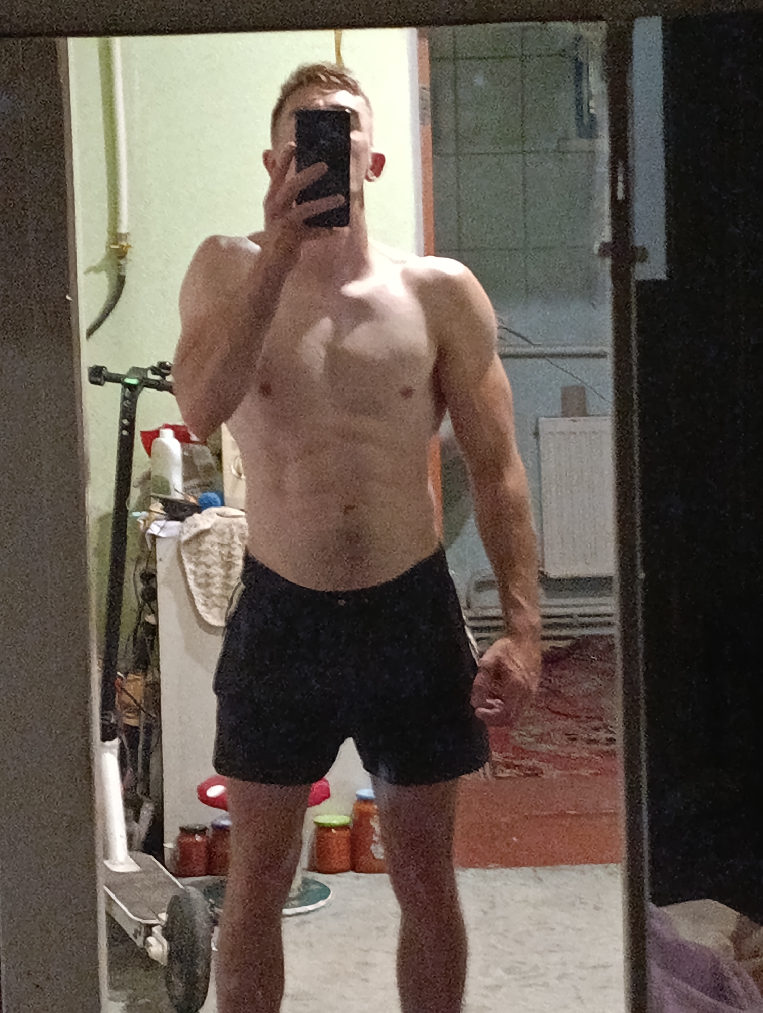 A man is standing in front of a mirror, taking a selfie. He is shirtless and wearing dark shorts. His hair is short and reddish-brown. He has abs and defined muscles. The background appears to be a room in an apartment or house, with some objects visible in the reflection of the mirror. To the left, there is a white scooter. To the right, there is a radiator and some jars on a shelf. The walls are painted a light green color. The lighting is dim. The image is grainy. The scene is consistent with an indoor residential setting.