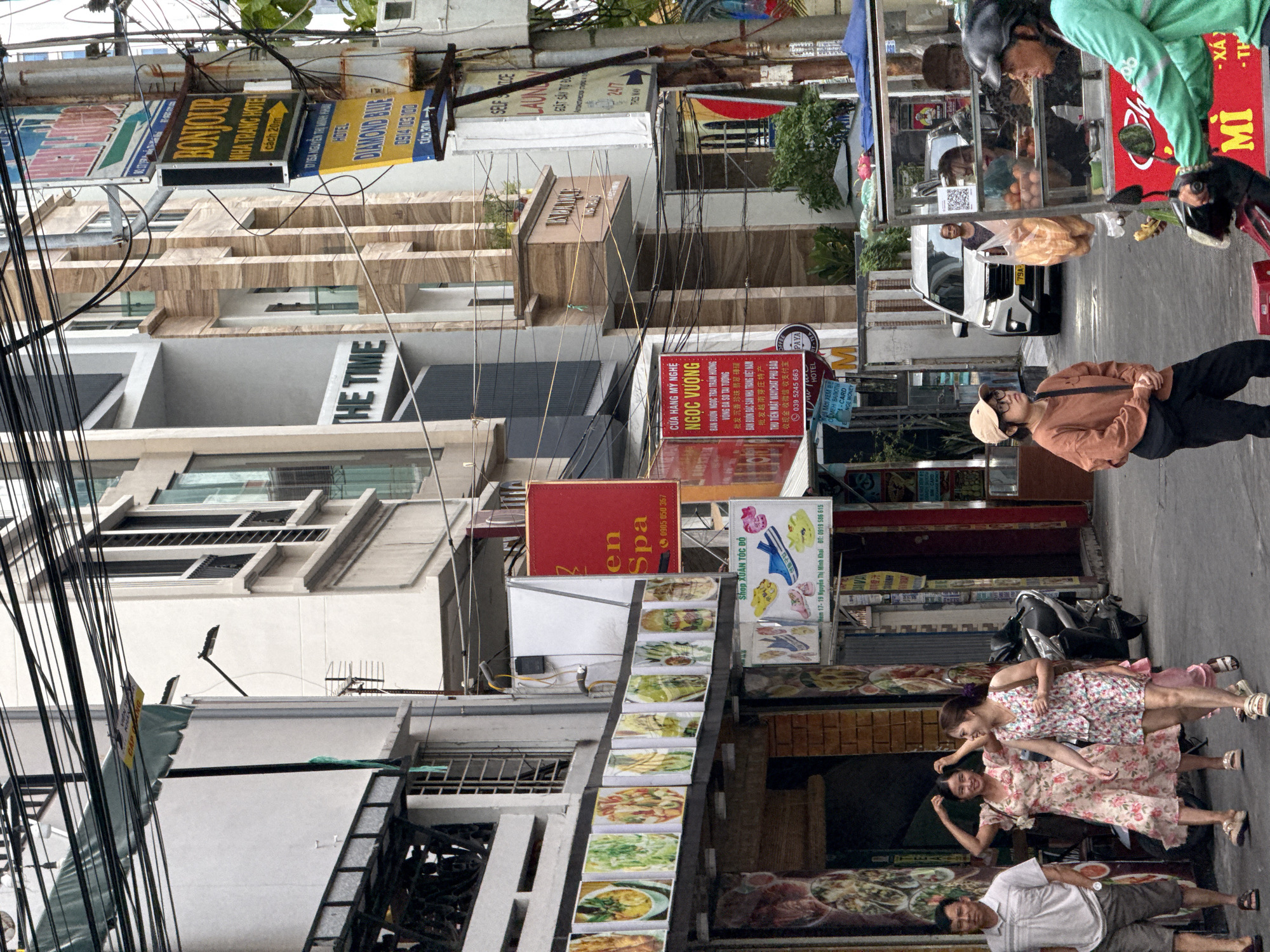 The image depicts a bustling street scene in Nha Trang, Vietnam. The street is lined with multi-story buildings adorned with various signs, some in Vietnamese, advertising hotels and businesses. Overhead, a complex network of electrical wires crisscrosses the sky.
In the foreground, several people are walking on the street. On the left, a man in a light-colored polo shirt and shorts walks with his hands in his pockets. Beside him, two women in floral dresses are walking and talking. The woman on the left has her hands raised as if adjusting her hair, and the woman on the right is smiling. Further down the street, a woman in a tan long-sleeved shirt and dark pants stands looking to her left.
To the right, a street vendor operates a cart filled with bread and other goods. The vendor, wearing a green top and a helmet, is on a scooter, interacting with someone behind the cart. A white SUV is parked further down the street.
The buildings showcase diverse architectural styles, with some featuring balconies and large windows. Signs for "BONJOUR NHA TRANG HOTEL" and "DIAMOND BLUE HOTEL" are visible, indicating tourist accommodations. Other signs advertise services like "EN SPA" and "LAUNDRY." Food stalls and shops are apparent along the street, displaying images of dishes. The overall impression is a vibrant urban environment with a mix of commercial activity and pedestrian traffic.