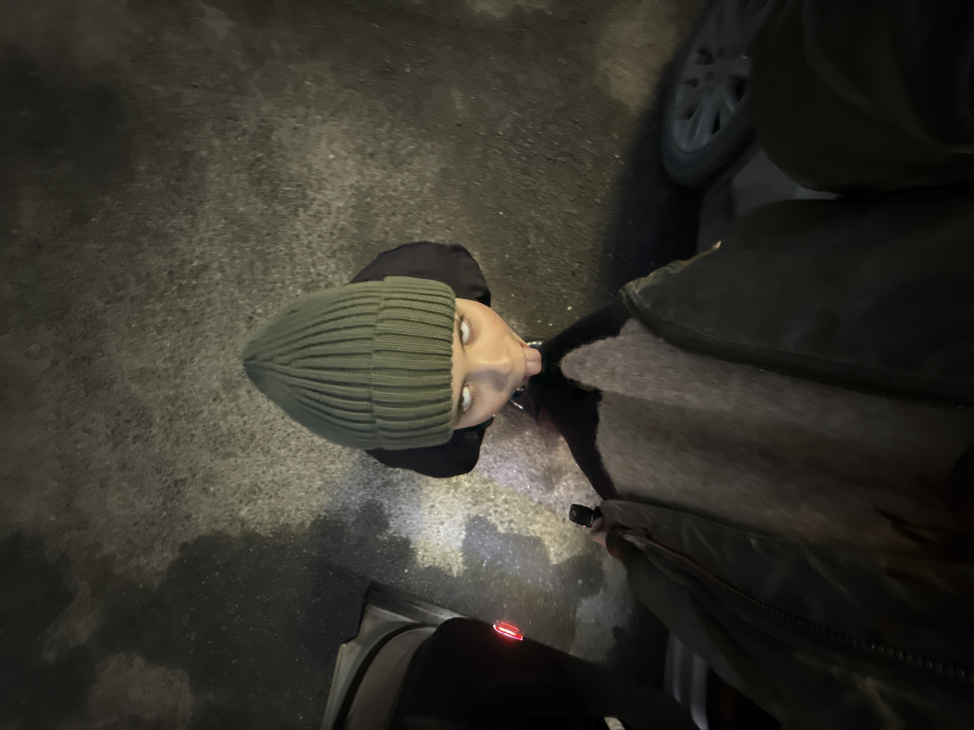 A person wearing a green beanie stands on a paved surface at night. The surrounding environment appears dark. Part of a car, including a wheel and an open door with a red light, is visible. The person wears a dark-colored jacket and sweater. The location is not determinable from the provided visual information.