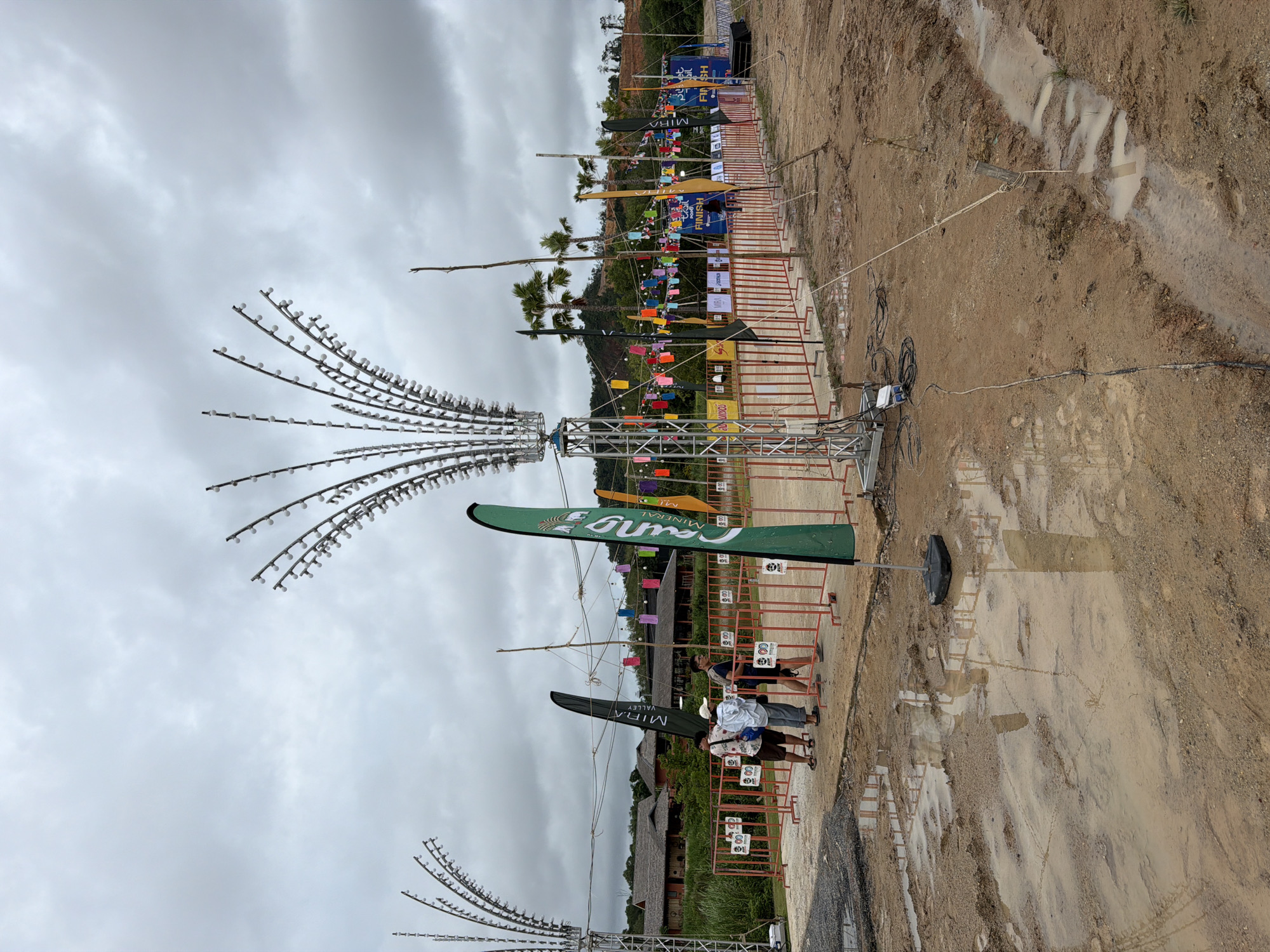 The image depicts an outdoor event or festival area under a cloudy sky in the city of Si Sunthon, Thailand. In the foreground, the ground is muddy with several large puddles, suggesting recent rainfall. A trench filled with muddy water runs along the right side of the frame.
Several people are visible near a red metal barricade. Three individuals, appearing to be attendees, stand together in casual clothing. Behind the barricade, there are various flags and banners, some with text and logos. Notable flags include a tall green flag with white text on the right, and a dark green flag with white text to the left of it.
Large metal structures with strings of lights resembling fireworks or decorative lamps are prominently featured on the left and right sides of the image. In the background, there are trees, some palm trees, and buildings with traditional-looking roofs. Colorful paper lanterns are strung across the area between poles, adding to the festive atmosphere.
The scene suggests a temporary setup for an event, possibly a race finish line or a public gathering, with a focus on decorations and crowd management. Electrical cables are visible on the ground, connecting to the lighting structures. The overall environment is somewhat dishevelled due to the muddy conditions, but elements of celebration are evident.
