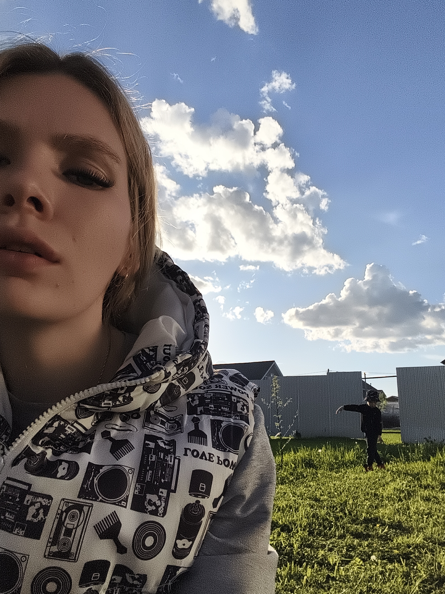 The image shows a young woman in the foreground, taking a selfie. She is wearing a hooded vest with a black and white pattern of music-related images like boomboxes and vinyl records. In the background, there's a yard with green grass. A person, likely a child, is standing in the grass, possibly gesturing or walking, wearing a dark outfit. A fence is visible behind the child, and beyond the fence, there's a glimpse of houses, suggesting a residential area. The sky is blue with puffy white clouds. The location appears to be Русский Вожой, Russia.