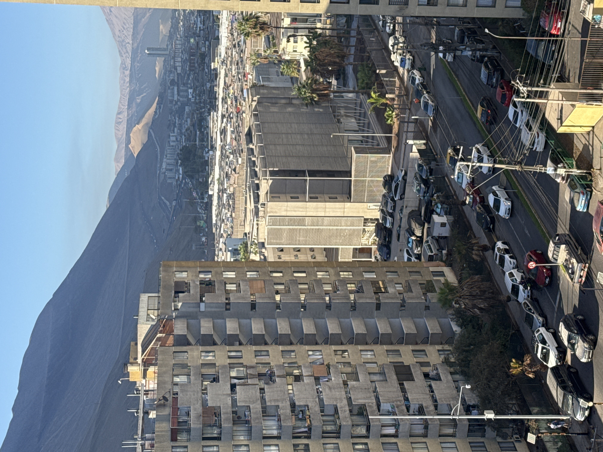 This image, captured from a high vantage point, presents a sprawling urban landscape backed by arid mountains under a clear blue sky. The photo is rotated clockwise by 90 degrees; when viewed upright, the sky is at the top and the ground at the bottom.
In the immediate foreground and to the left, a tall, multi-story apartment building with a distinctive Brutalist architectural style stands prominently. It features numerous recessed concrete balconies and windows, giving it a complex, textured facade.
Below and to the right of this building, a busy street scene unfolds. Multiple lanes of road are visible, lined with numerous parked cars of various models and colors (predominantly white, grey, black, and some red). Several utility poles, burdened with a dense network of overhead wires, run alongside the street. Patches of greenery and palm trees are interspersed along the roadside and between buildings.
Further into the middle ground, the city expands into a dense conglomeration of buildings, indicative of a populous urban center. In the far background, large, rugged mountains with a desert-like appearance rise sharply, forming a dramatic natural backdrop to the urban development. One crop shows a solitary tall building standing out amidst the distant cityscape and mountains.
The scene is bathed in bright sunlight, evidenced by the sharp shadows cast by buildings and vehicles, suggesting a late morning or early afternoon. The sky is a cloudless, vibrant blue. A few small human figures can be seen on the street or balconies, but they are not central to the composition. Visible text on street signs includes a red circular "No Entry" sign and a white 'N' in a red circle, likely indicating "No Parking."