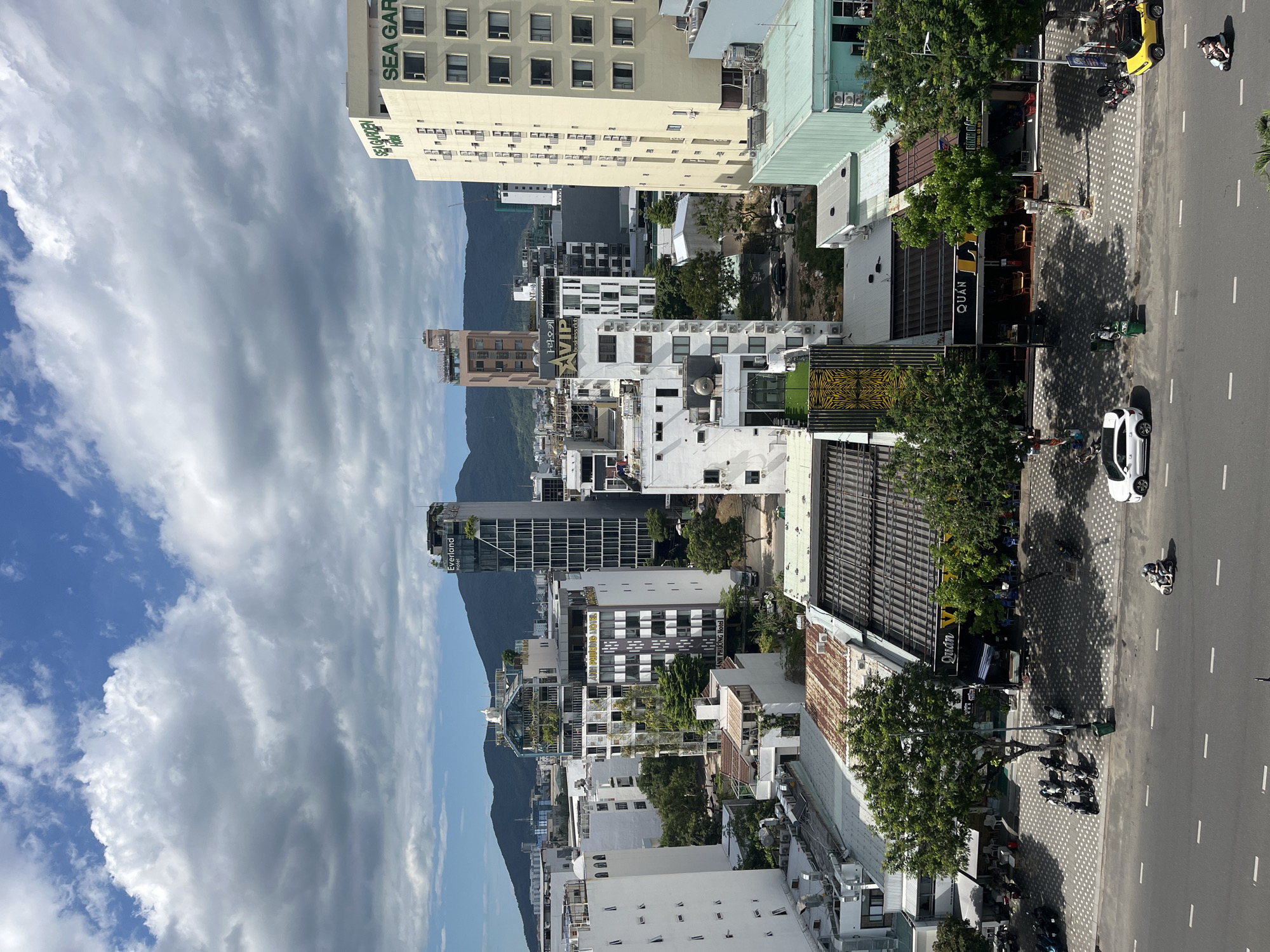In this aerial photograph, a city skyline is presented under a partly cloudy sky. The foreground features a street with a white car and several motorcycles, some of which are parked along the sidewalk. Buildings of various heights and architectural styles dominate the midground and background, with some featuring signage for hotels and businesses such as "SEA GARDEN Hotel" and "ANH PHUONG HOTEL." Trees are interspersed among the buildings, providing splashes of green. In the distance, mountains form a natural backdrop to the urban landscape. The overall scene captures a bustling city environment with a blend of modern architecture and natural elements.