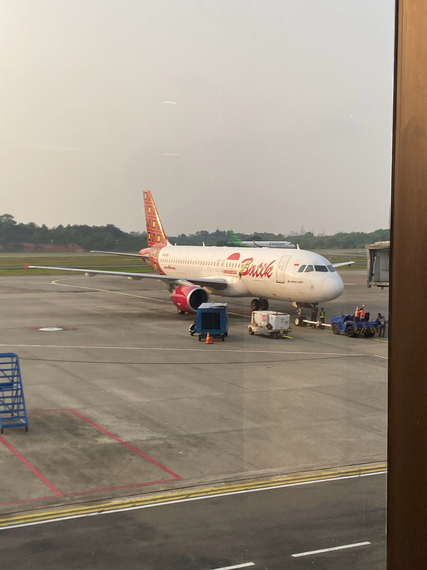 An airplane with "Batik" branding is parked at an airport gate. The airplane is white with red and orange accents on its tail and engines. Ground crew are visible near the plane, and a jet bridge is connected to the aircraft. Another airplane with "Citilink" branding is visible in the background. The scene appears to be an airport tarmac, with planes and ground support equipment. The image seems to be taken from inside a terminal, looking out at the aircraft. The weather appears overcast.
