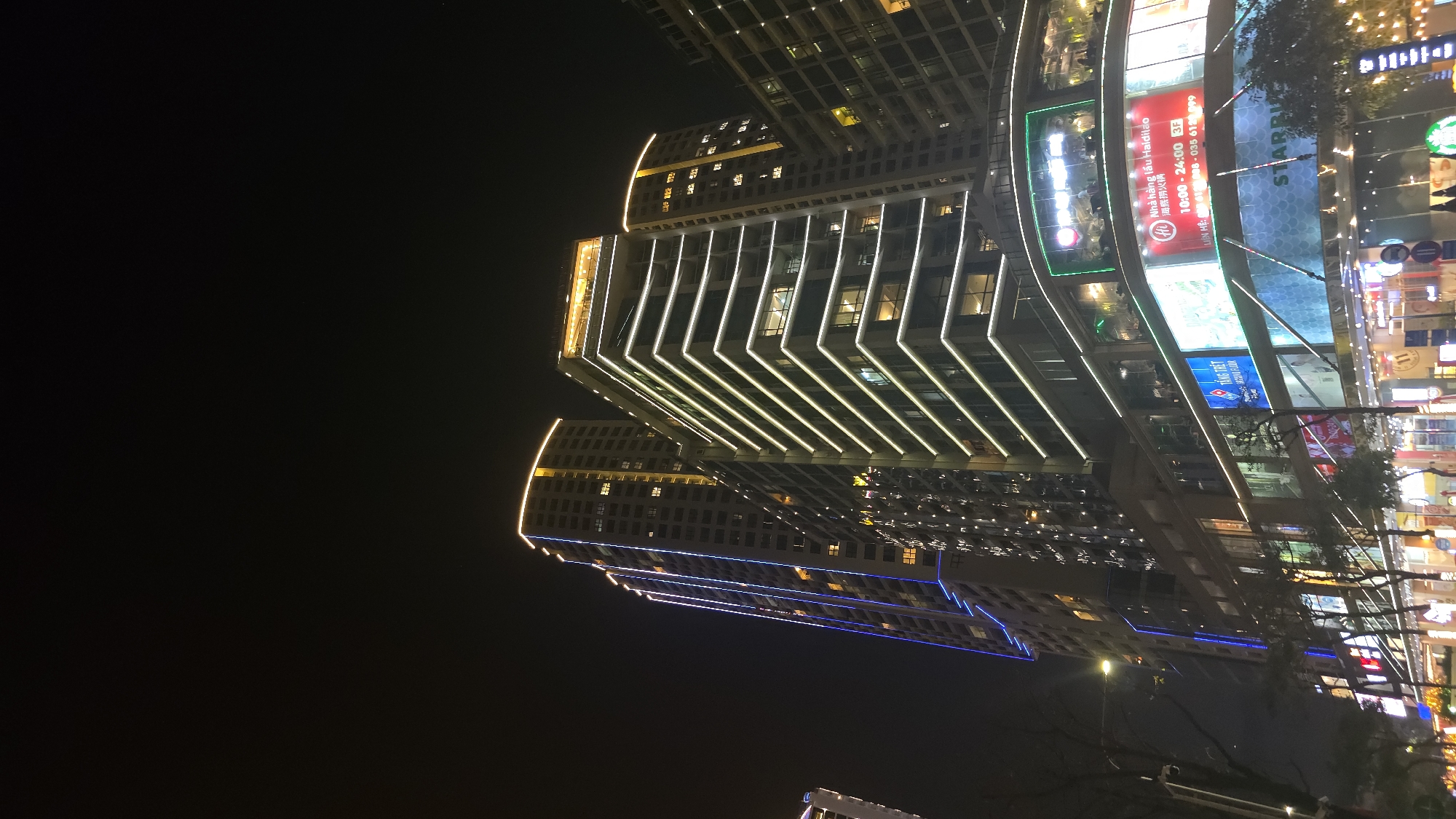 The image depicts a nighttime urban scene in Nha Trang, Vietnam, characterized by multiple illuminated high-rise buildings. The primary focus is a modern commercial and residential complex featuring a multi-story base with retail and dining establishments and tall residential or hotel towers above.
The most prominent building on the right features numerous horizontal white LED light strips outlining each floor, with a golden-lit top section. To its left, another taller skyscraper is adorned with vertical blue LED light strips running along its facade, also topped with golden illumination. A third, partially visible building is to the far left. The sky overhead is dark, indicating night.
The lower levels of the complex host various commercial businesses. A large red sign prominently displays "Nha hàng Haidilao" (Haidilao Restaurant), with operating hours "10:00 - 24:00" and "3F" indicating its location on the third floor. Below this, a large "STARBUCKS" sign is visible. To the left of Haidilao, a blue sign mentions "Domino's Pizza" and "Tầng Trệt" (Ground Floor). Other signs with Vietnamese text and brand logos are visible throughout the commercial base, suggesting a bustling shopping and dining area.
Small figures of people are discernible in the brightly lit, open-air sections of the commercial complex, particularly on the upper dining terraces, indicating active patronage. At street level, obscured by trees and the lower part of the buildings, faint outlines of individuals are visible, suggesting pedestrian activity. Streetlights and sparse tree branches are present along the base of the buildings, bordering what appears to be a street or pedestrian walkway. The overall scene conveys a vibrant, modern city environment after dark.