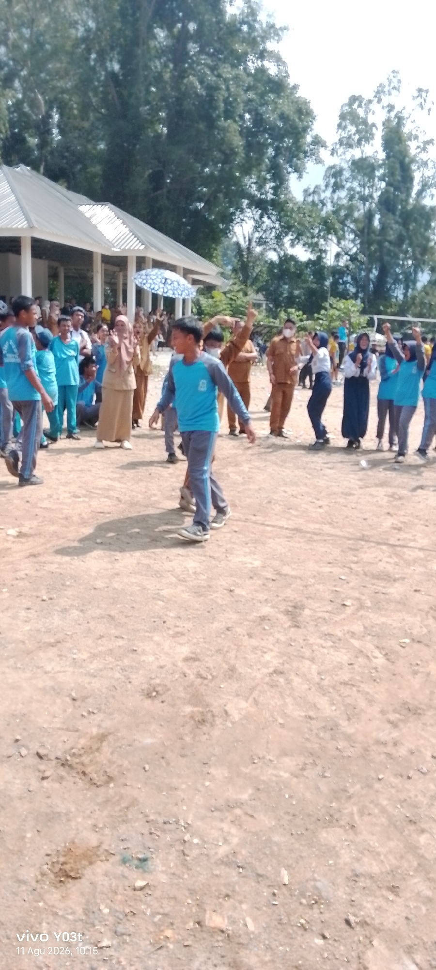 In the city of Tirawuta, Indonesia, a group of students and adults are gathered outdoors, seemingly for an event. In the foreground, a young male wearing a blue shirt and grey pants with blue stripes is walking towards the right of the frame, looking ahead with a slight smile. Behind him, another young male in a similar outfit is seen from the side, also walking. A group of people in the middle ground are watching or participating in the activity. Some are wearing tan uniforms, possibly teachers or officials, while others are in blue shirts and headscarves, appearing to be students. One person in a tan hijab is clapping, and another holding a patterned umbrella stands nearby. In the background, more individuals are visible, and a building with a white roof and open sides is present. The ground is dusty and appears to be a sports field or open area. The overall scene suggests a casual, outdoor gathering possibly related to a school event or competition.