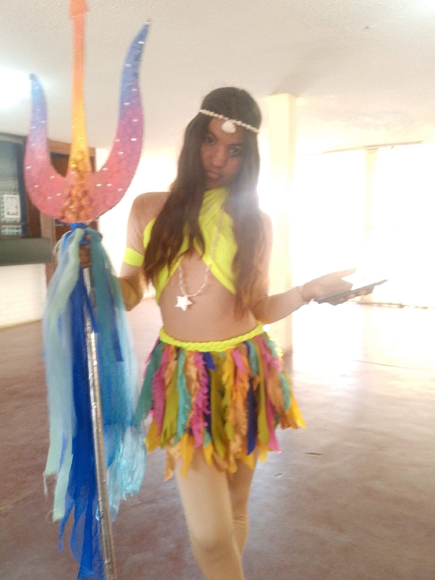 A woman is standing indoors, holding a large, decorated trident in her left hand. She is wearing a yellow crop top with tan sleeves, a colorful feather skirt, and tan leggings. A pearl-like headband adorns her hair, and she wears a star-shaped necklace. Her right hand holds a dark object, possibly a phone or wallet. The trident is decorated with blue, pink, and yellow hues and adorned with flowing blue fabric. The background is bright, suggesting a well-lit interior space with neutral-colored walls. The location is likely an indoor space, potentially in the city of Juan Griego, Venezuela, although this cannot be confirmed solely from the image.