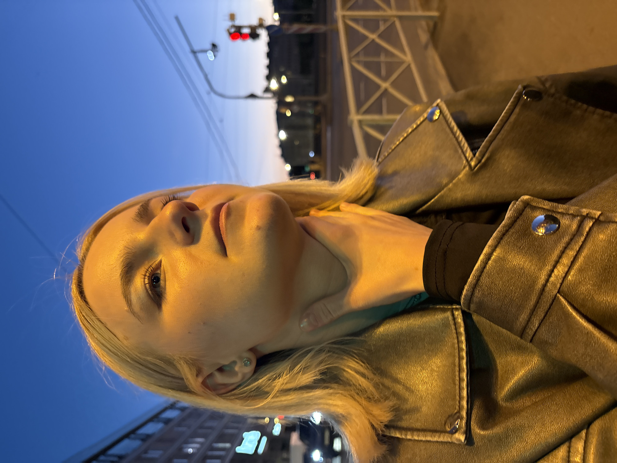 A blonde woman with light blue eyes is shown from the chest up, standing outdoors at dusk in Saint Petersburg, Russia. She is wearing a metallic leather jacket over a black garment. Her right hand is resting on her throat, and she is looking upwards. The background shows a city street with a traffic light showing red, streetlights, buildings, and power lines against a clear blue sky. A metal railing is visible in the foreground.