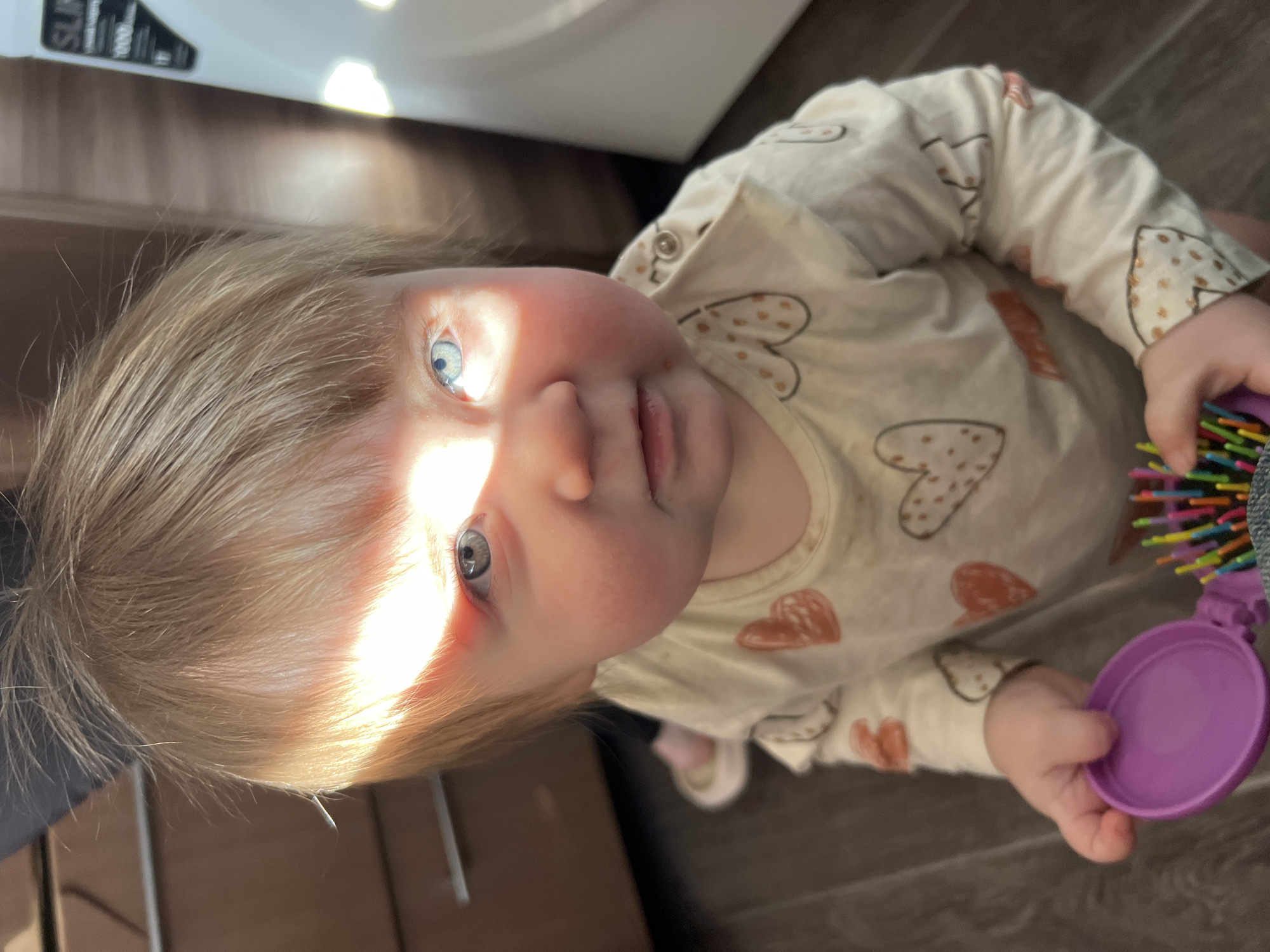A baby with blonde hair and blue eyes is looking up at the camera. The baby is wearing a long-sleeved shirt with hearts on it and holding a purple toy with colorful bristles. The baby's face is partially illuminated by a bright light. The background is blurred, but appears to show a wooden floor and a white appliance. The location is unknown and there is no confirmation of it being Chaikovsky, Russia.