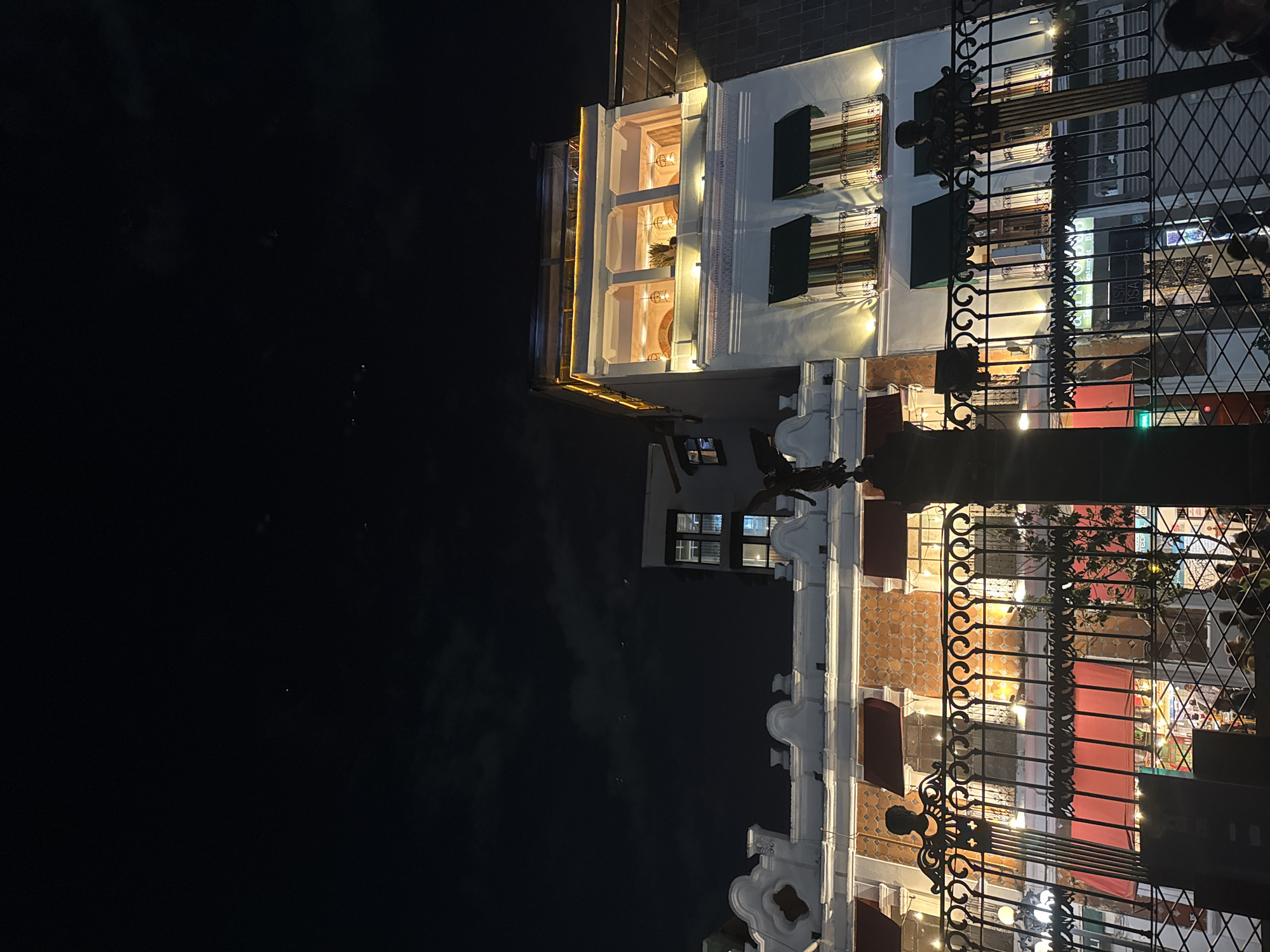 The image shows a building at night in Puebla, Mexico. The building has multiple stories with lit windows. Some windows have awnings. An ornate wrought-iron fence is in the foreground. The sky is dark.