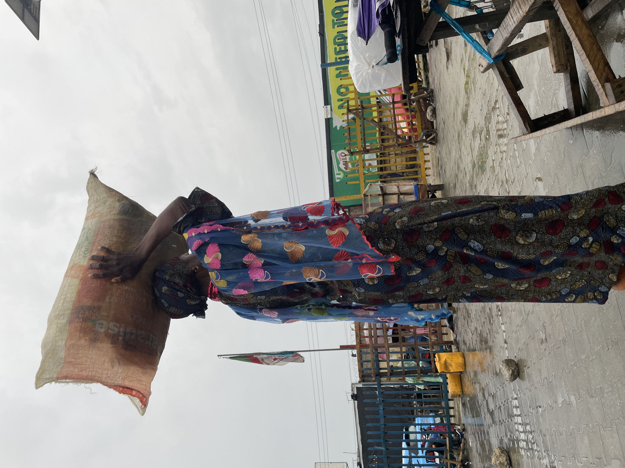 A woman in Lagos, Nigeria carries a large sack on her head in an outdoor market-like setting. She is wearing a patterned dress with a blue shawl adorned with red and pink designs. Her head is wrapped with a patterned cloth. The sack, which appears to be made of burlap, is hoisted on her head. In the background, there are stalls with goods and signs. A sign with the words "NO NEED TO FEAR" is visible, along with a Milo advertisement. The ground is wet, suggesting it has recently rained. A Nigerian flag waves in the background. The overall scene suggests a bustling, commercial environment.