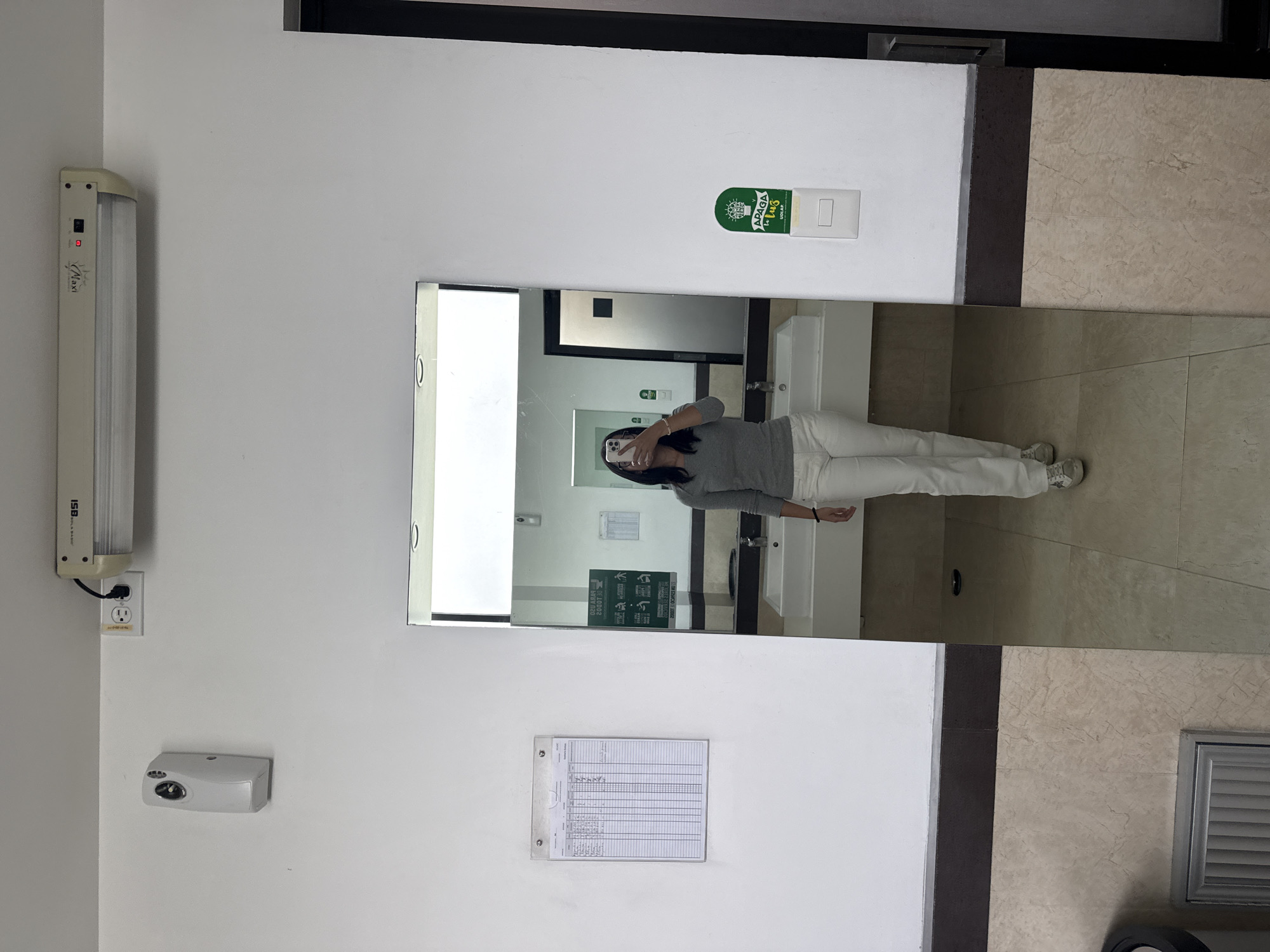 A person stands in the reflection of a mirror in a bathroom in San Andrés Cholula, Mexico. The individual, clad in a gray shirt and white pants, is holding a phone in front of their face, presumably taking a photo. The bathroom features white walls, a marble floor, a sink, and various fixtures. These fixtures include a fluorescent light fixture, an electrical outlet, a soap dispenser, a mirror, a checklist on a clipboard, and a light switch reminder sticker.
