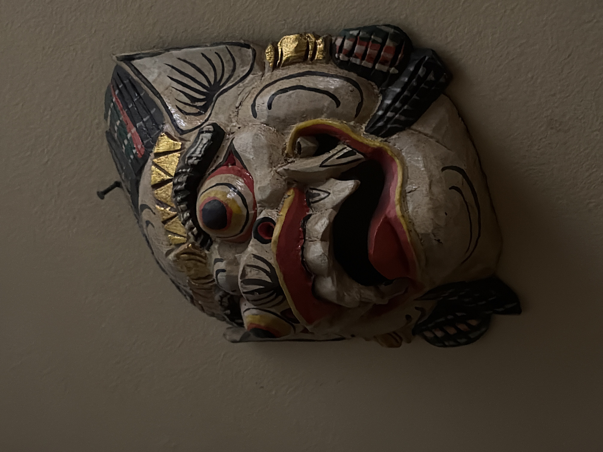 The image displays a close-up, dimly lit view of an ornate, carved wooden mask affixed to a plain, light-colored wall. The mask is predominantly off-white or light beige, adorned with intricate painted details. Its most prominent features include large, circular eyes with concentric rings of red, yellow, and black, and a wide-open mouth revealing visible teeth and a dark, red-lined interior, possibly representing a tongue or throat.
Stylized black lines delineate eyebrows, facial contours, and decorative patterns across the mask's surface. Gold accents are visible around the crown of the head and near the cheeks, adding a decorative flourish. Additional colors, including red and dark green, are incorporated into patterned sections on the sides of the mask, which appear to be stylized ears or decorative extensions. The mask's texture suggests carved wood.
A small, dark nail or hook is visible on the upper-left side of the mask, indicating its mounting method. The background is a uniform, subtly textured wall, likely indoor. The low lighting casts significant shadows, particularly on the right side and lower parts of the mask, enhancing its three-dimensional form and creating a dramatic atmosphere. No activity is depicted, and no text is visible in the frame.