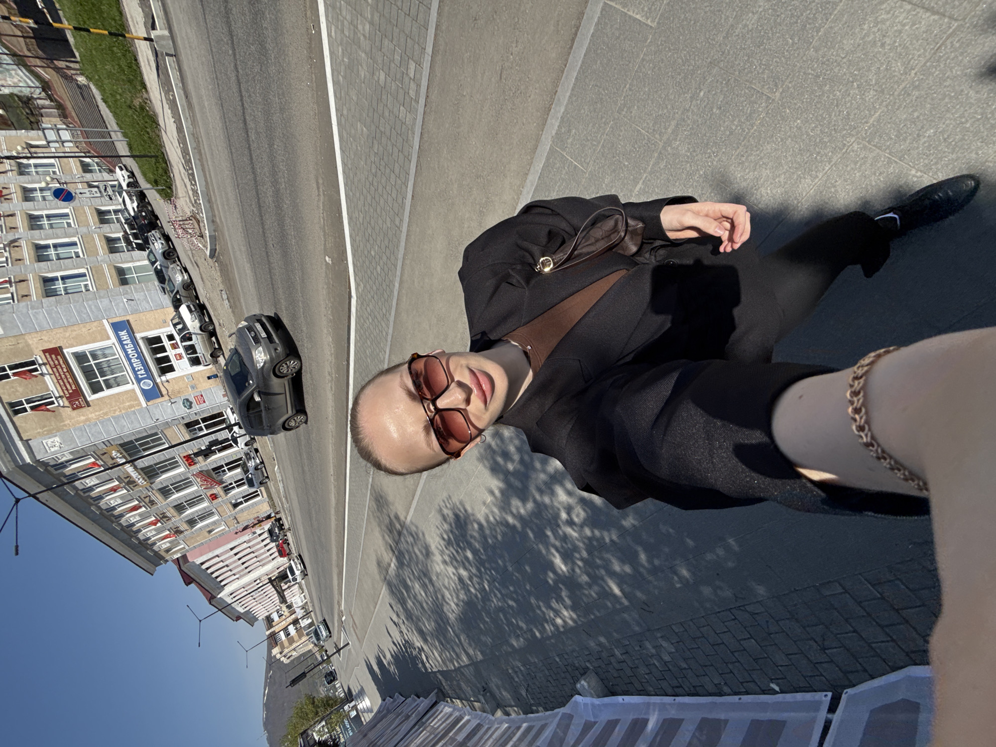 A woman takes a selfie in Petropavlovsk-Kamchatsky, Russia, on a sunny day. She is standing on a sidewalk next to a street with parked cars and a multi-story building with a "Gazprombank" sign. The woman is wearing sunglasses, a brown shirt, and a black blazer. She is holding a small brown purse. In the background, the building has several windows and red flags hanging. The sidewalk is paved with grey stones and has tree shadows cast upon it. The street is asphalt and has white lane markings.