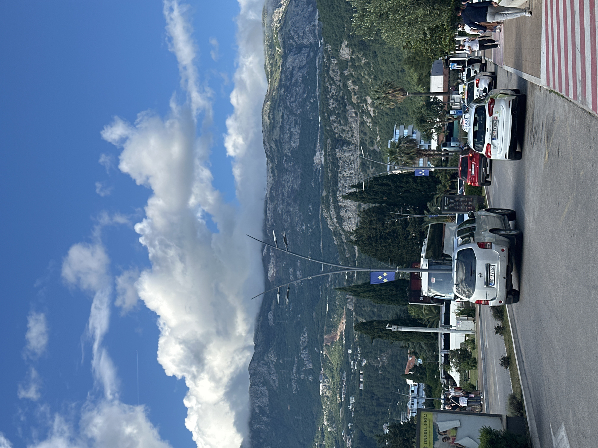 The image captures a sunny daytime scene in Budva, Montenegro, with a scenic backdrop of rugged, green mountains partially veiled by clouds. A bright blue sky with scattered white cumulus clouds, and a faint airplane contrail, stretches above the peaks.
In the foreground and midground, a paved road is bustling with activity. Numerous cars are present, with several prominent white vehicles identified as taxis by "TAXI" signs on their roofs. Visible license plates, such as "BD TX004" on a white car and "BD AY451" on a silver SUV, indicate their origin in Budva. A red car is also seen on the road.
To the right of the road, a pedestrian zone features distinct red and white striped markings, where several people are walking or standing. Lush green vegetation, including palm trees, lines the roadside, alongside buildings nestled into the foothills. A flagpole displays a blue flag adorned with yellow stars, similar to the European Union flag. On the far left, a large billboard is partially visible, displaying the text "ANABEL ARTO". The overall impression is that of a vibrant and scenic town on a clear day.