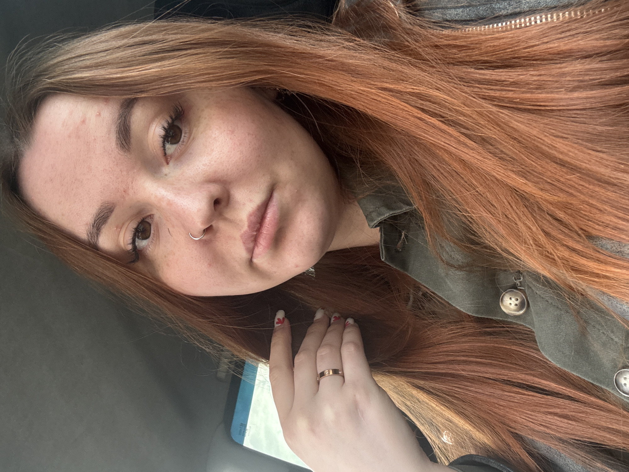 A selfie of a fair-skinned woman with light brown eyes, freckles, and red-brown hair is positioned at a slight angle. She is wearing a nose ring and a rose gold ring with diamonds on her ring finger. Her nails are painted white with small red designs. She is wearing a green jacket with brass buttons and a zipper. The background suggests she is inside a vehicle, possibly a car, with a gray interior visible. The location is not discernible as being in the city of Chaikovsky, Russia.