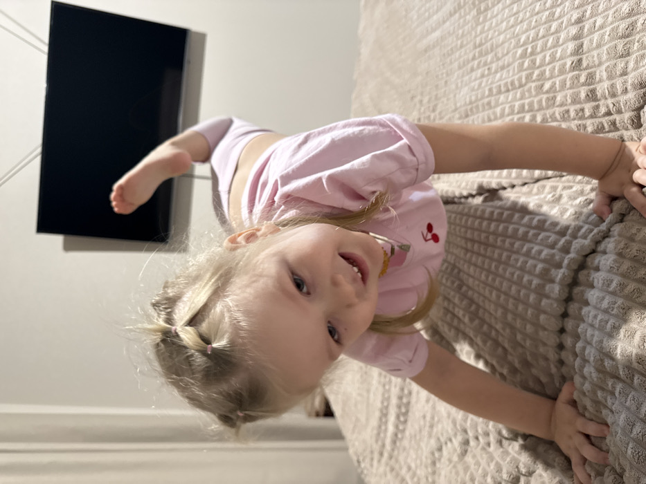A young girl in a pink shirt is pictured upside down, gripping the edge of a light-colored textured bedspread. She has blonde hair styled in pigtails, and her facial expression is a smile. A black television is mounted on a light gray wall in the background, with a bare foot also visible.