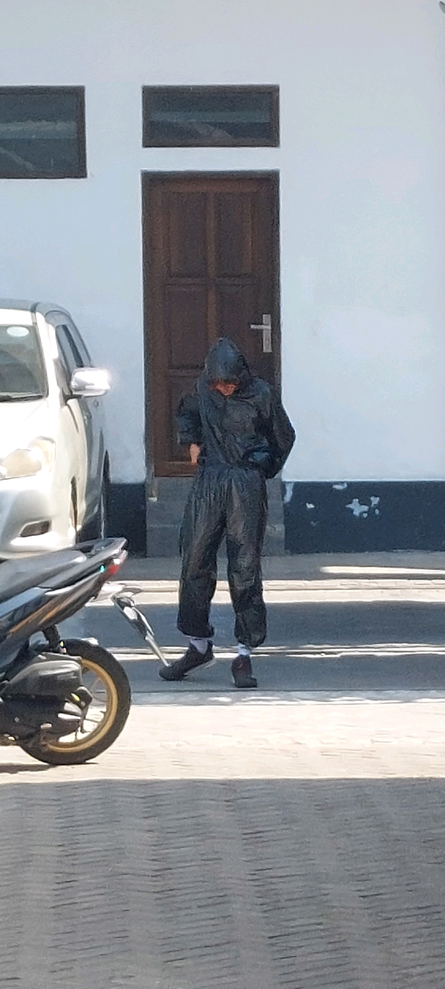 A person wearing a dark, hooded, possibly disposable, protective suit stands in front of a brown wooden door with a silver handle. The suit appears to be gathered at the ankles and wrists. The person is also wearing dark shoes and white socks. A silver car is parked to the left of the person. A black motorcycle is parked to the left and slightly in front of the car. The scene is outdoors, with a light-colored building and a dark, textured surface in the foreground. The lighting suggests it is daytime. The overall setting appears to be an urban or suburban area.